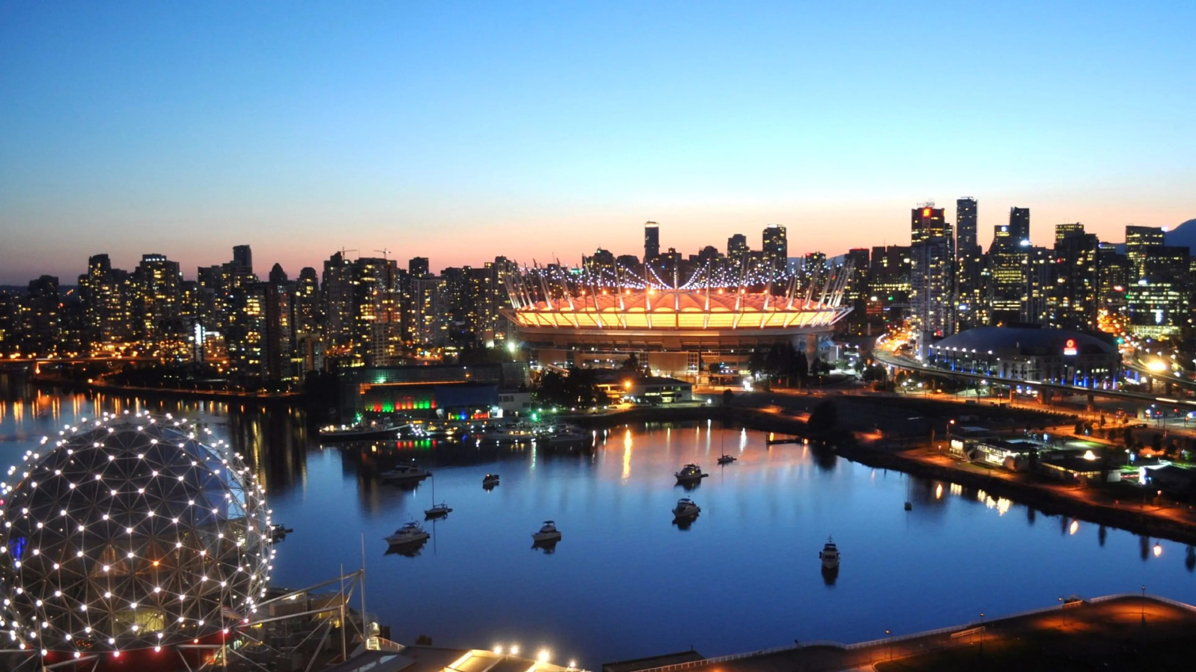 City skyline at dusk with illuminated BC Place stadium, Science World, and boats on calm water.