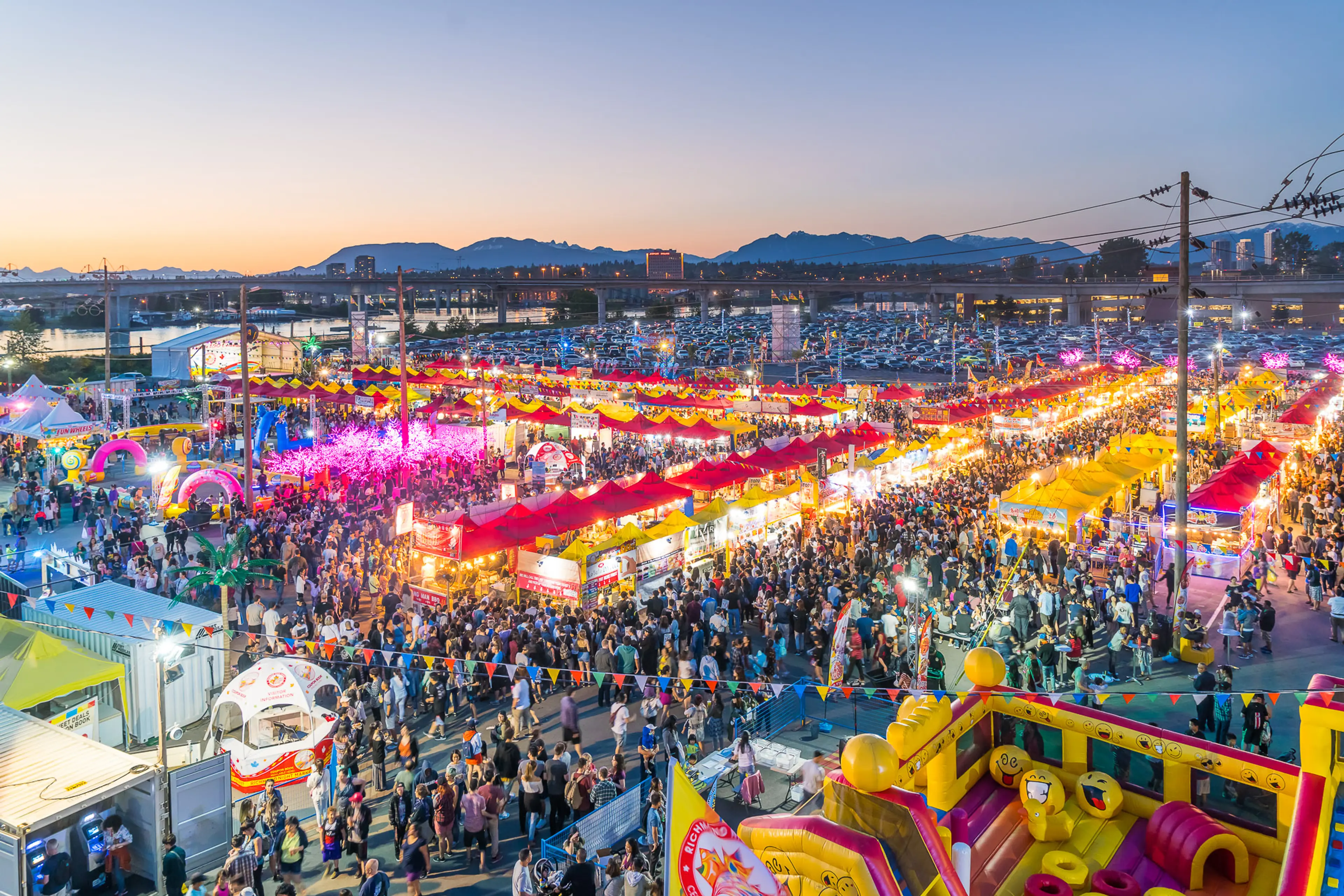 Crowded outdoor festival at dusk with colorful tents, bright lights, and an inflatable play area in the foreground at the Richmond Night Market..