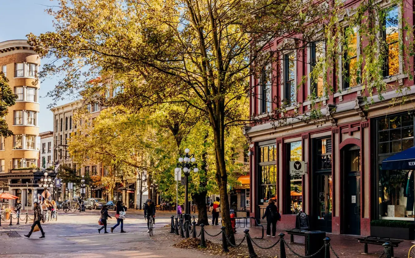 A lively street scene in historic Gastown featuring a classic red brick building, vintage globe streetlamps, and pedestrians walking along a textured cobblestone street shaded by autumn trees.
