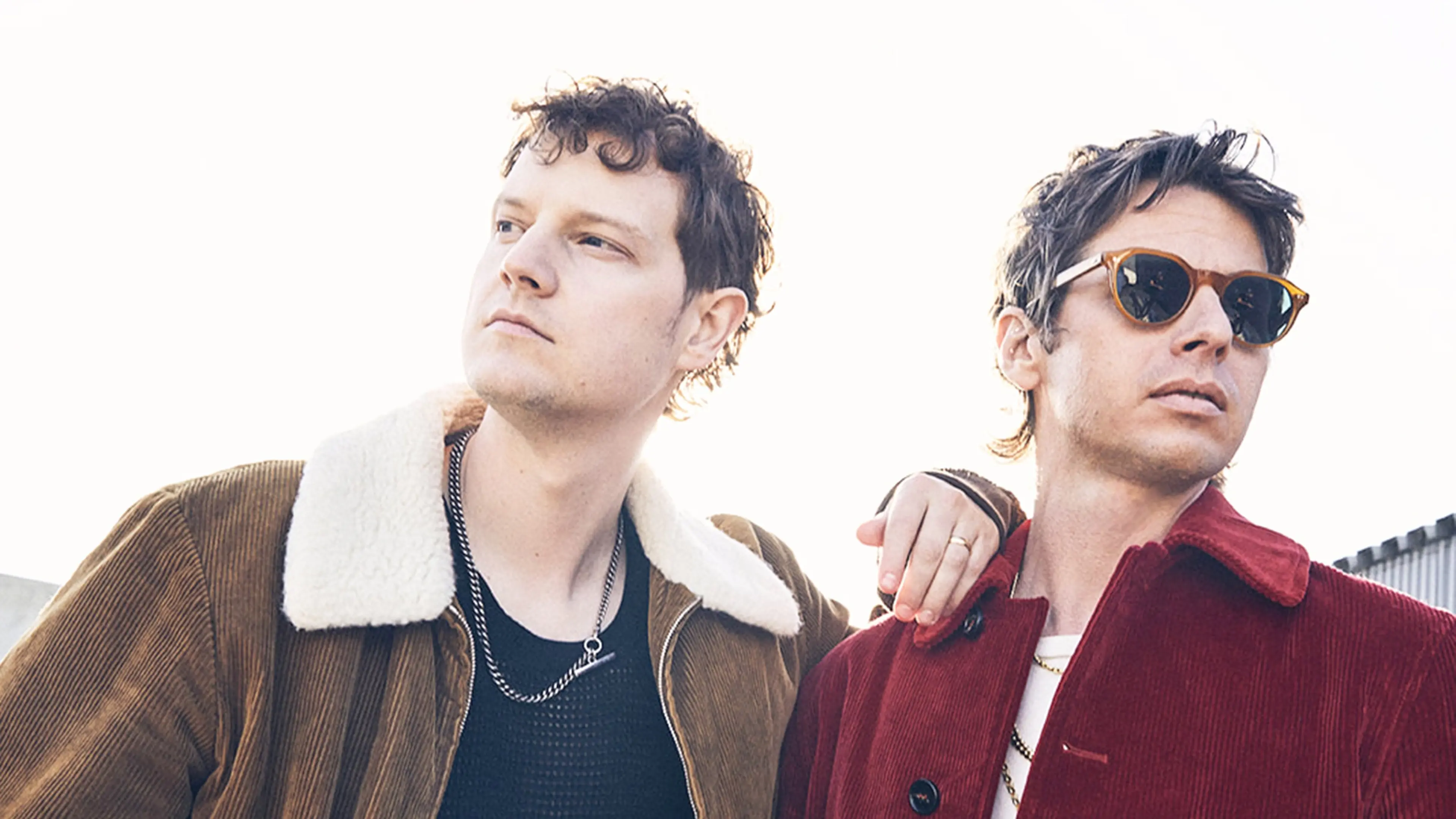 Two people wearing corduroy jackets, one brown with a white collar and the other red with sunglasses, looking away from the camera.
