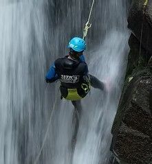 Person wearing a blue helmet and black gear rappelling down a waterfall beside a rocky cliff.