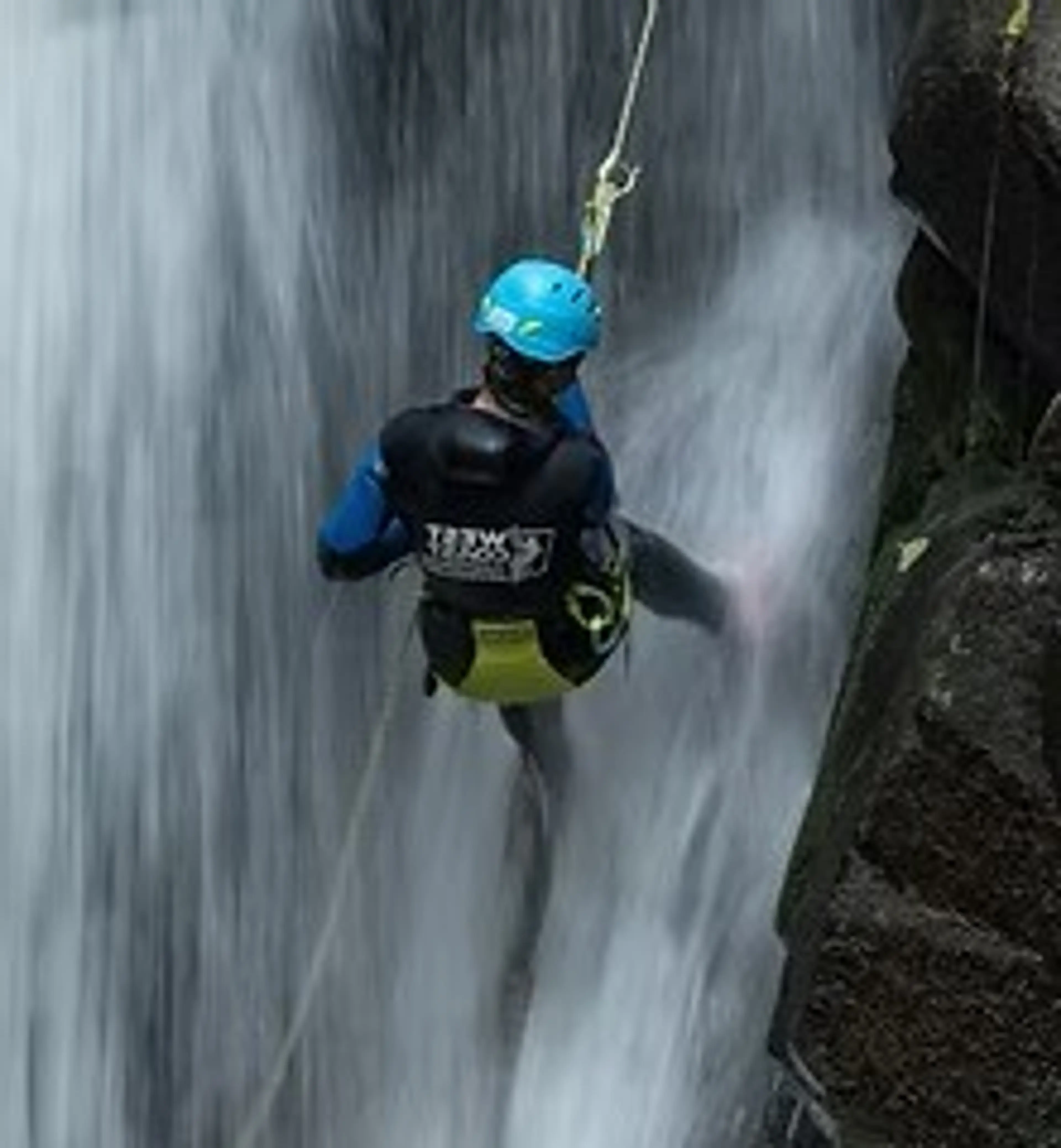 Person wearing a blue helmet and black gear rappelling down a waterfall beside a rocky cliff.