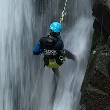 Person wearing a blue helmet and black gear rappelling down a waterfall beside a rocky cliff.