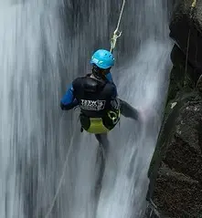 Person wearing a blue helmet and black gear rappelling down a waterfall beside a rocky cliff.