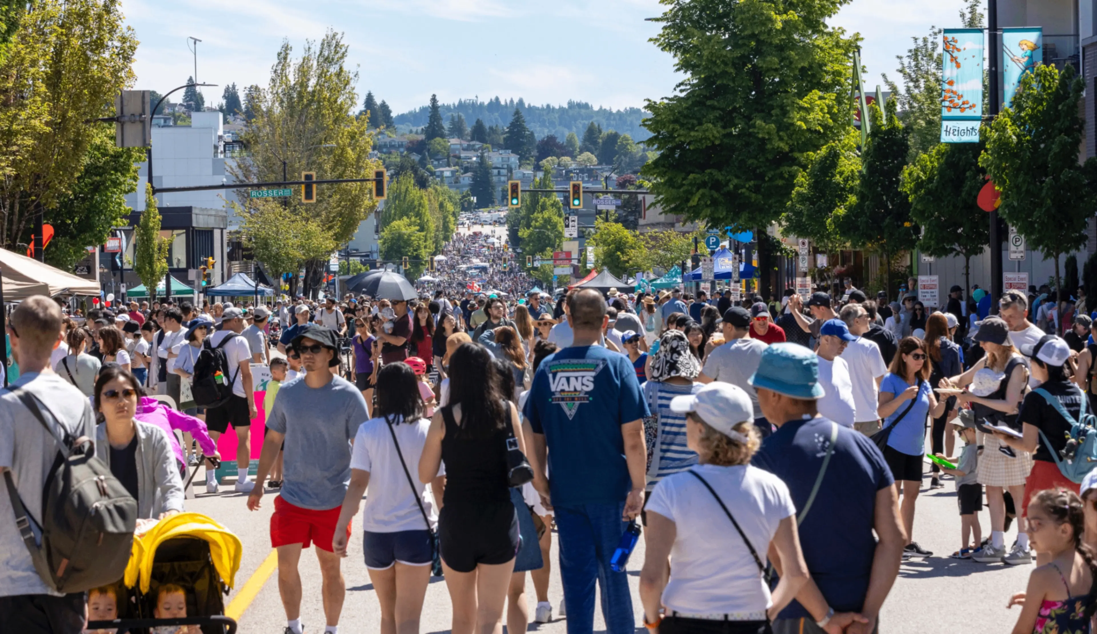 Crowded street festival with many people walking, some pushing strollers, under sunny weather with trees lining the street.