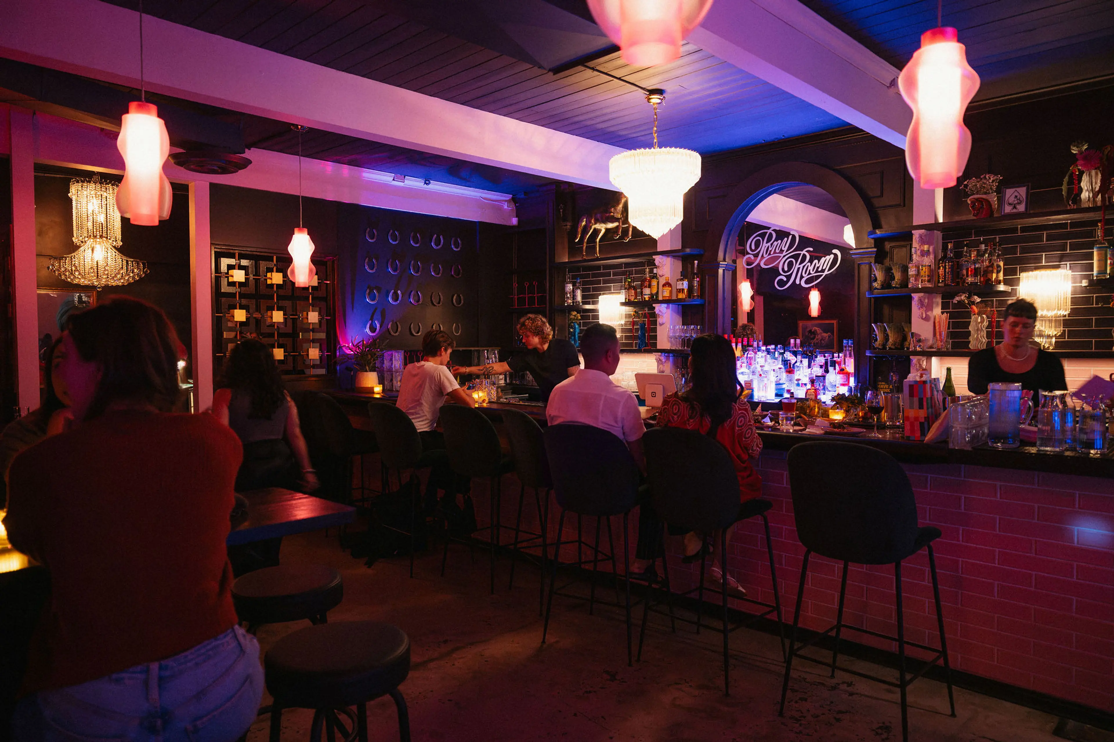 Dimly lit bar with people sitting on stools at the counter and tables, illuminated by pink and purple hanging lights.