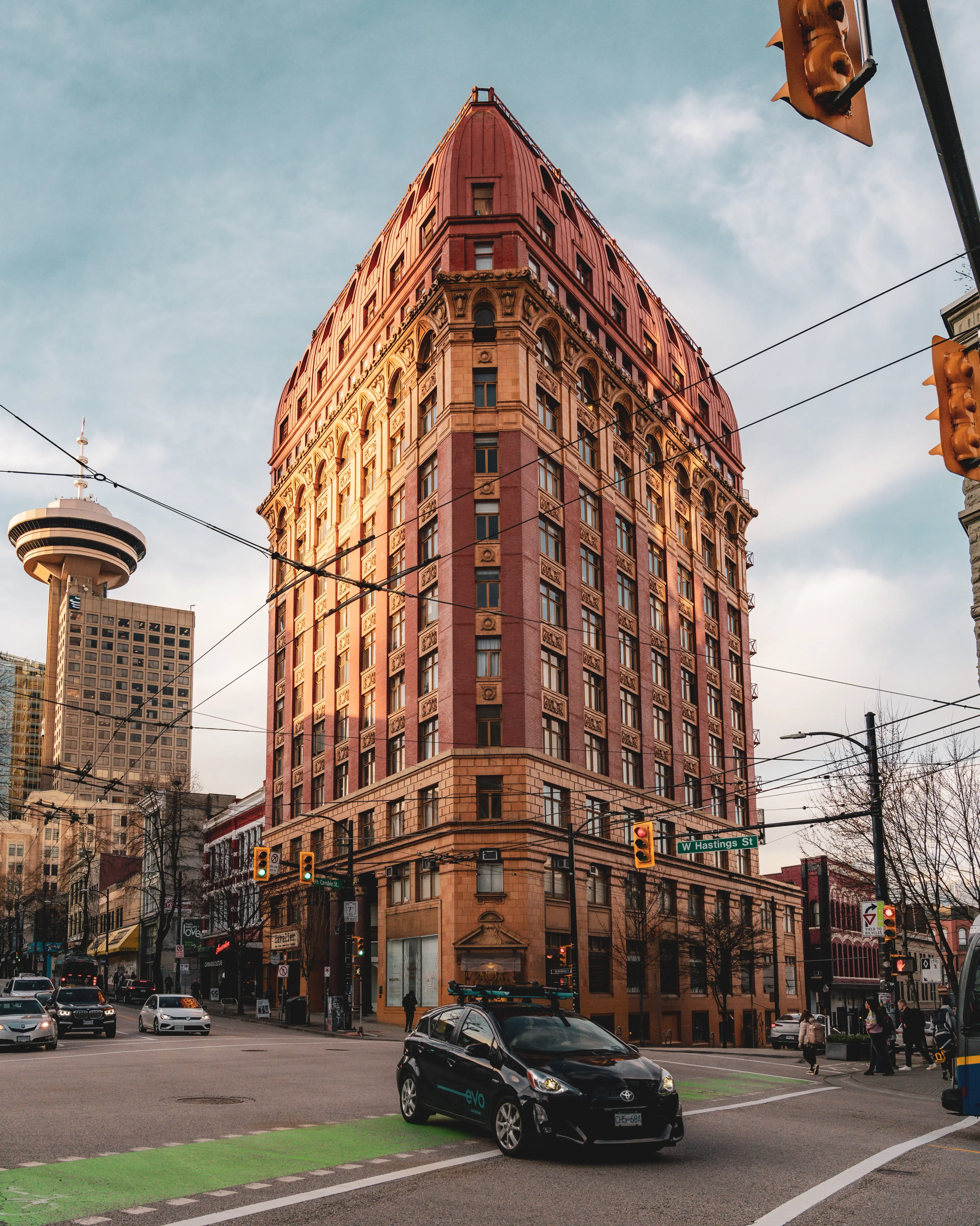 Tall historic building with red and beige facade at a busy city intersection with cars and pedestrians.