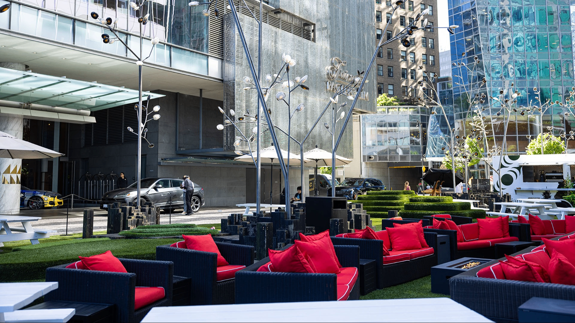 Outdoor seating area with black wicker sofas and red cushions on artificial grass in front of a modern building with glass and metal facade.