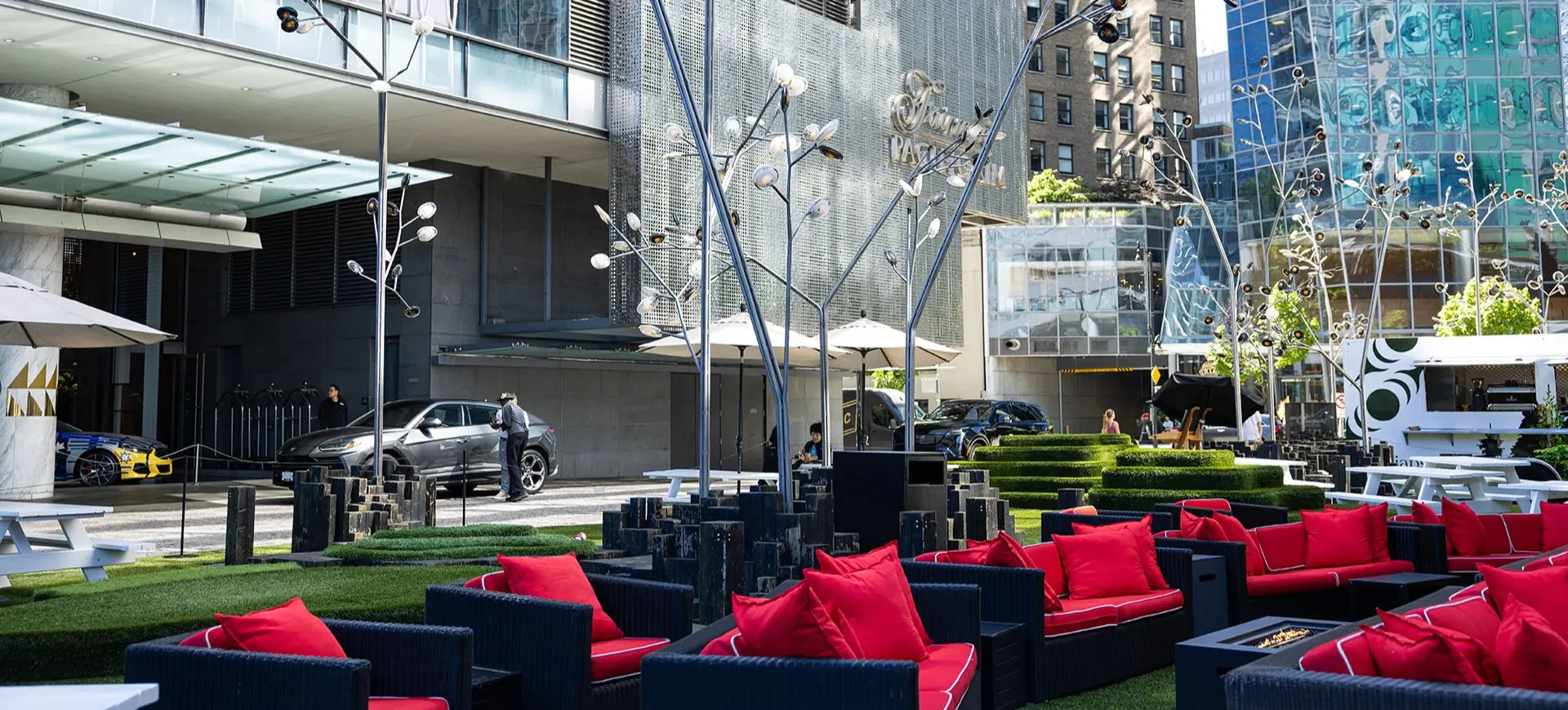 Outdoor seating area with black wicker sofas and red cushions on artificial grass in front of a modern building with glass and metal facade.