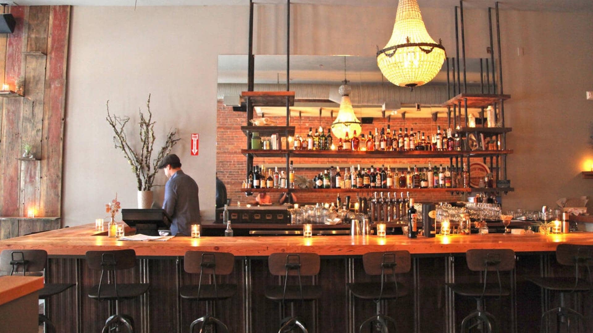 A bar tender is setting up a warmly lit modern bar with candles and a chandelier.