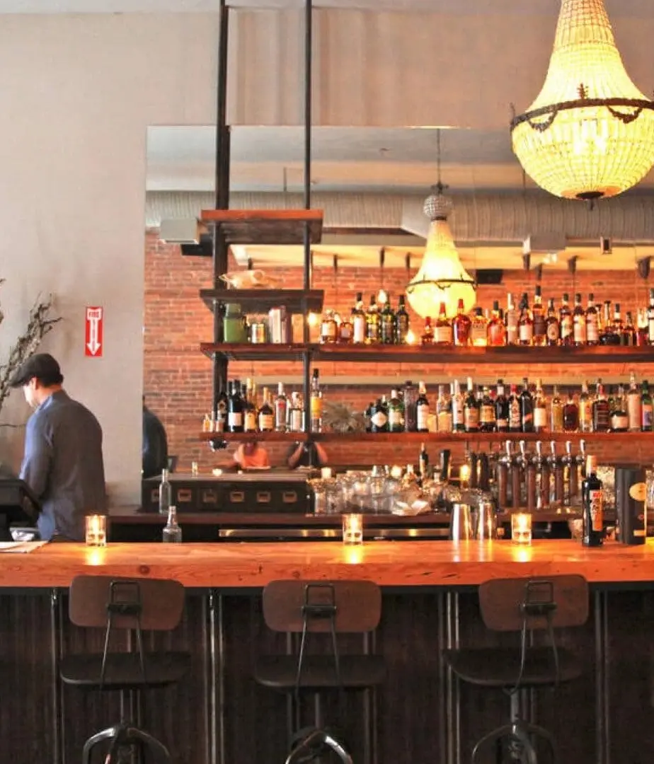 A bar tender is setting up a warmly lit modern bar with candles and a chandelier.