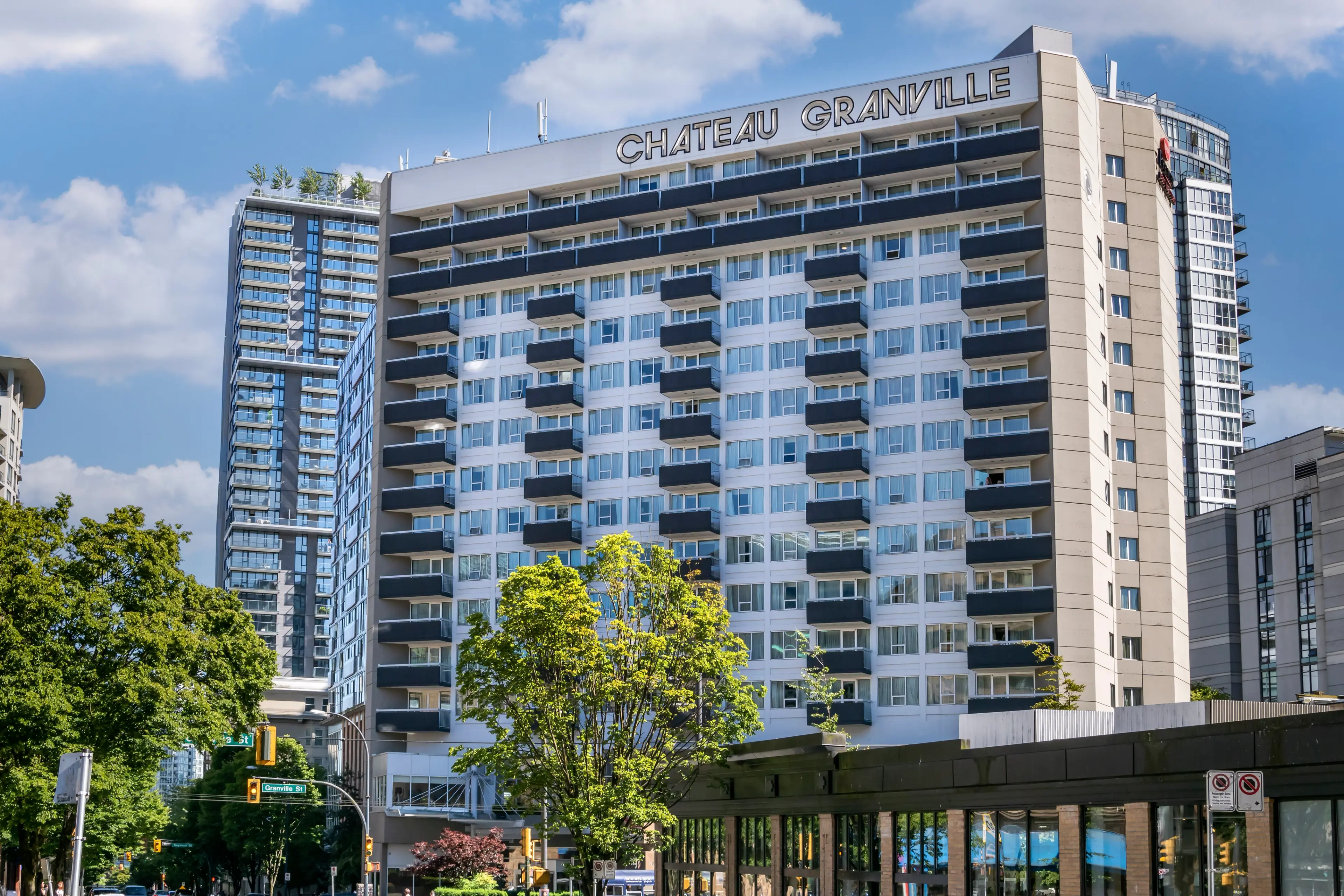 Tall building with balconies and sign reading Chateau Granville under a blue sky with clouds, surrounded by trees and other buildings.
