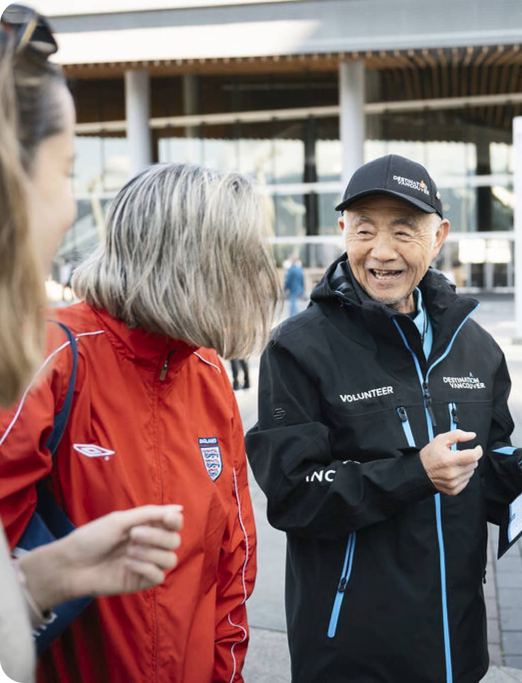 Three people outdoors, one wearing a red England jacket, another in a black volunteer jacket and cap, smiling and talking.