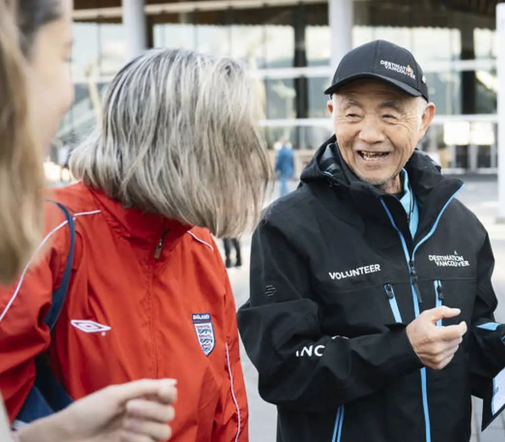 Three people outdoors, one wearing a red England jacket, another in a black volunteer jacket and cap, smiling and talking.