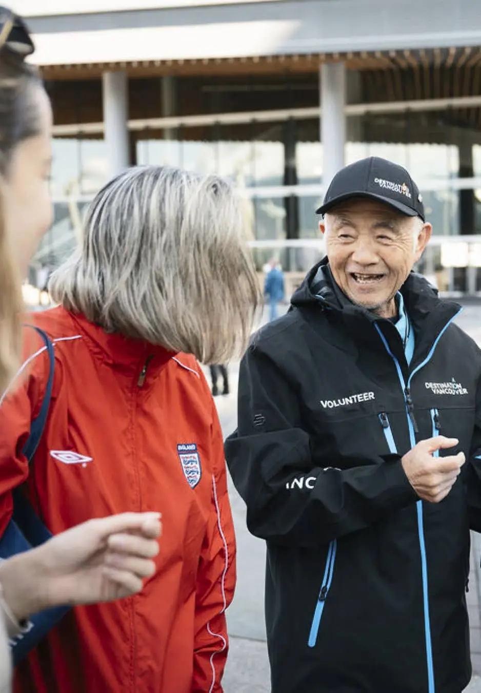 Three people outdoors, one wearing a red England jacket, another in a black volunteer jacket and cap, smiling and talking.