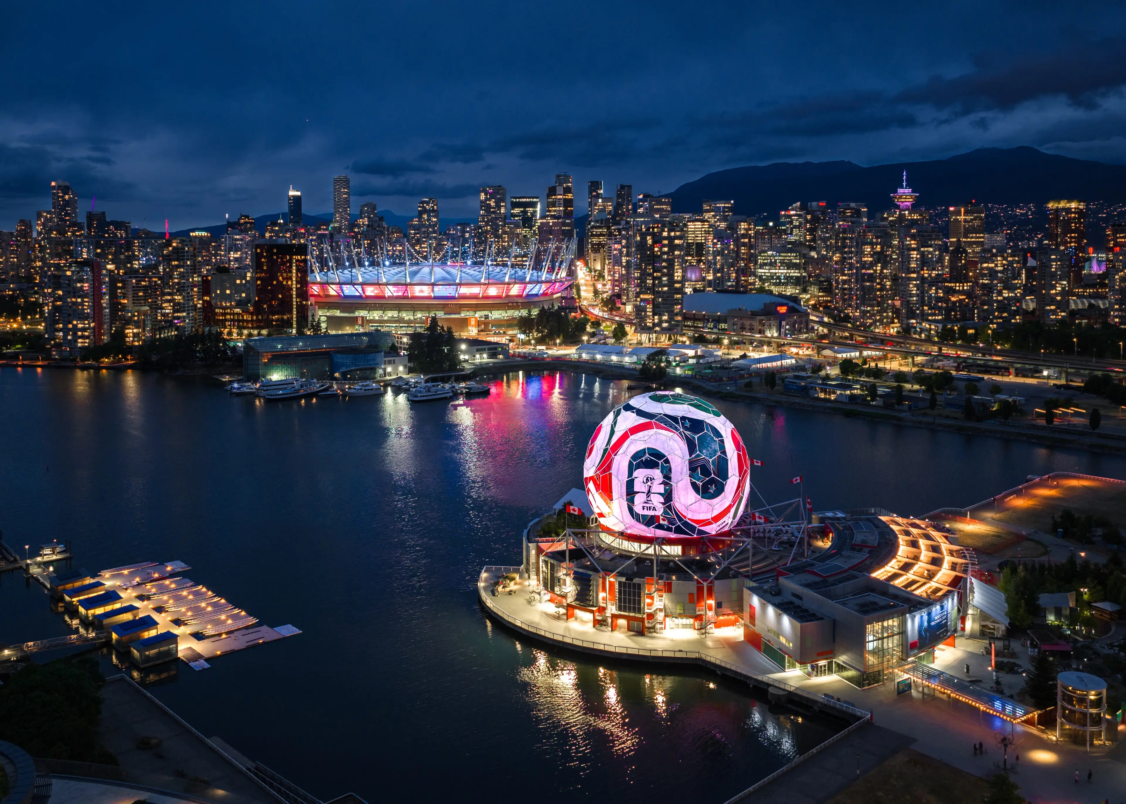 Night aerial view of a city skyline with a large illuminated spherical structure displaying a FIFA logo near the waterfront.