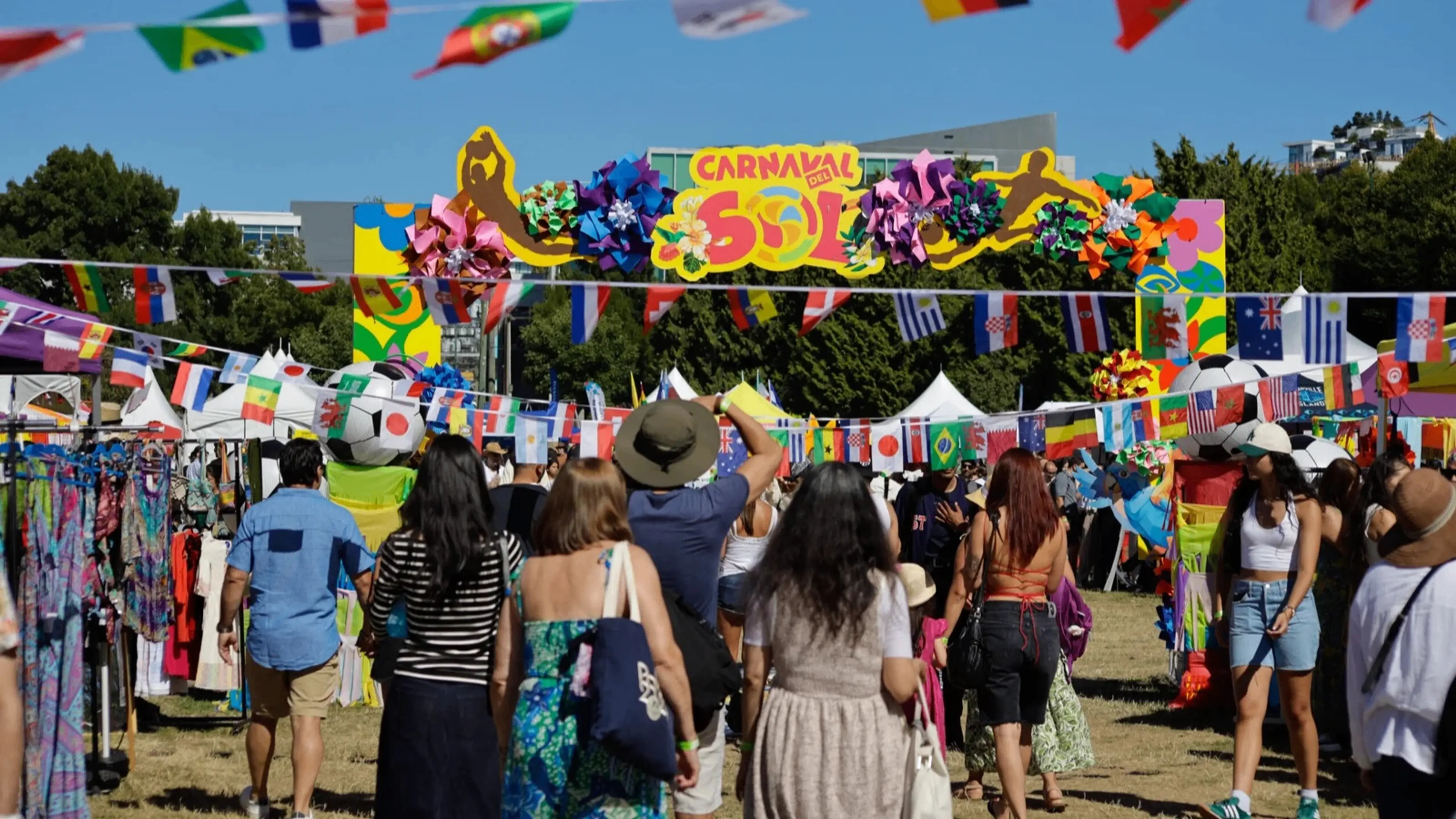 People walking under colorful flags and a sign reading Carnaval del Sol at an outdoor festival with vendor tents.