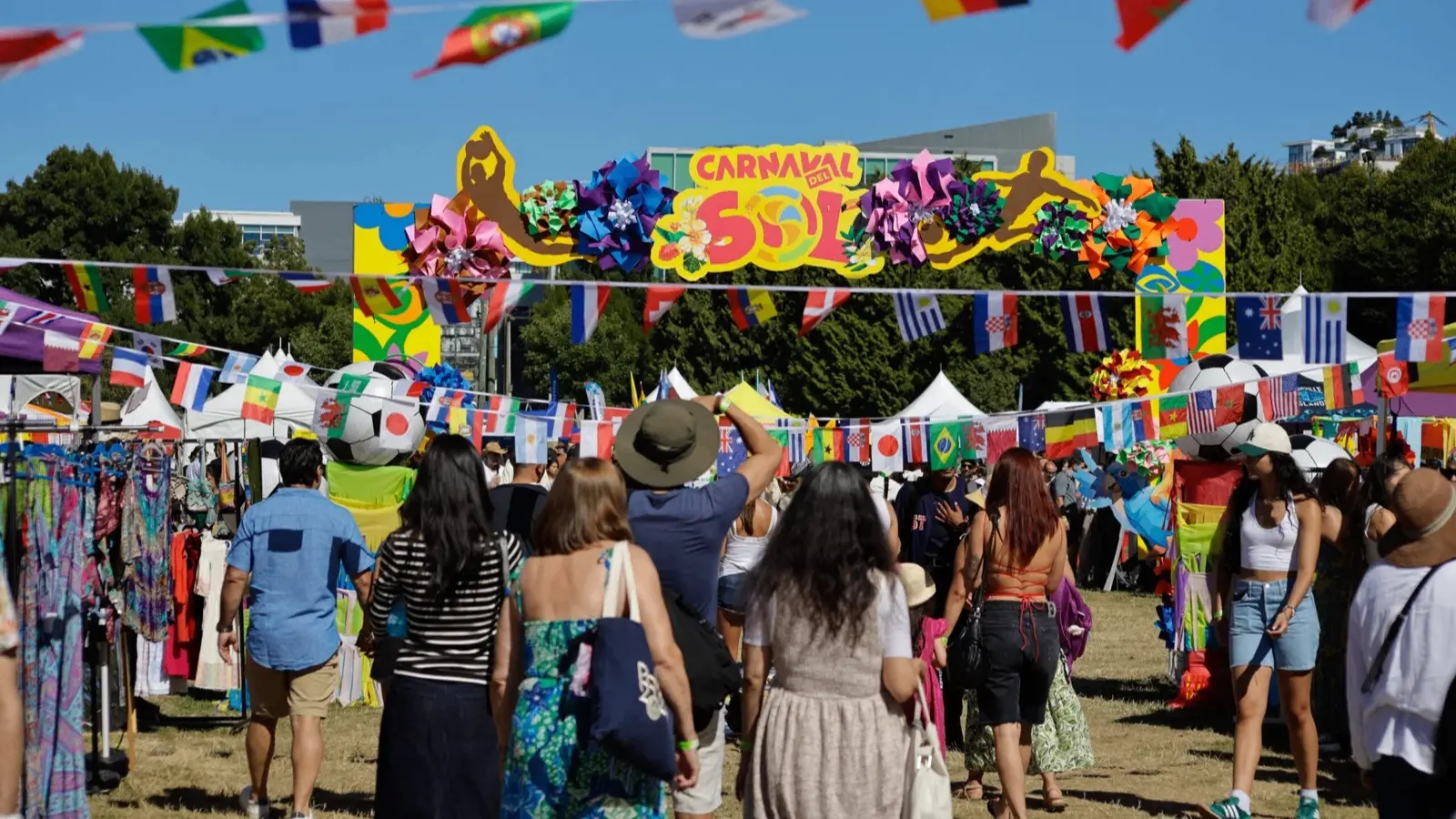 People walking under colorful flags and a sign reading Carnaval del Sol at an outdoor festival with vendor tents.