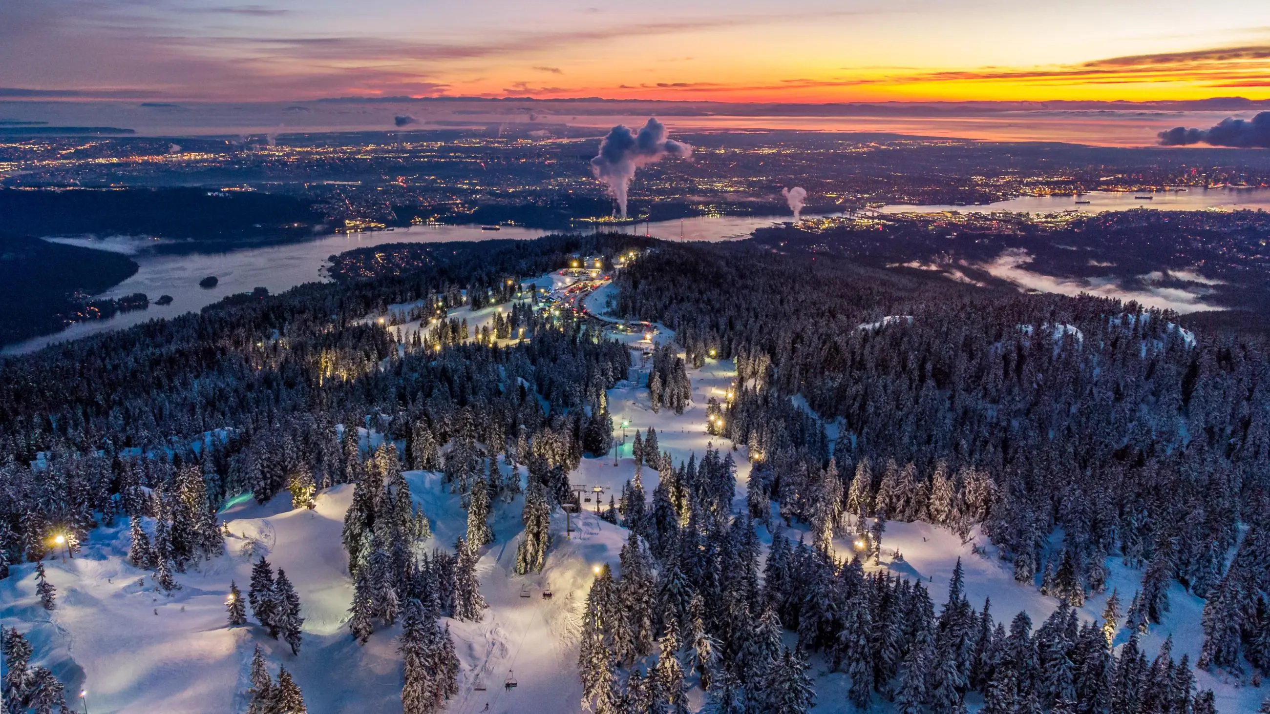 An aerial view of the ski runs at Mount Seymour resort lit up at night with the lights of Vancouver in the background.