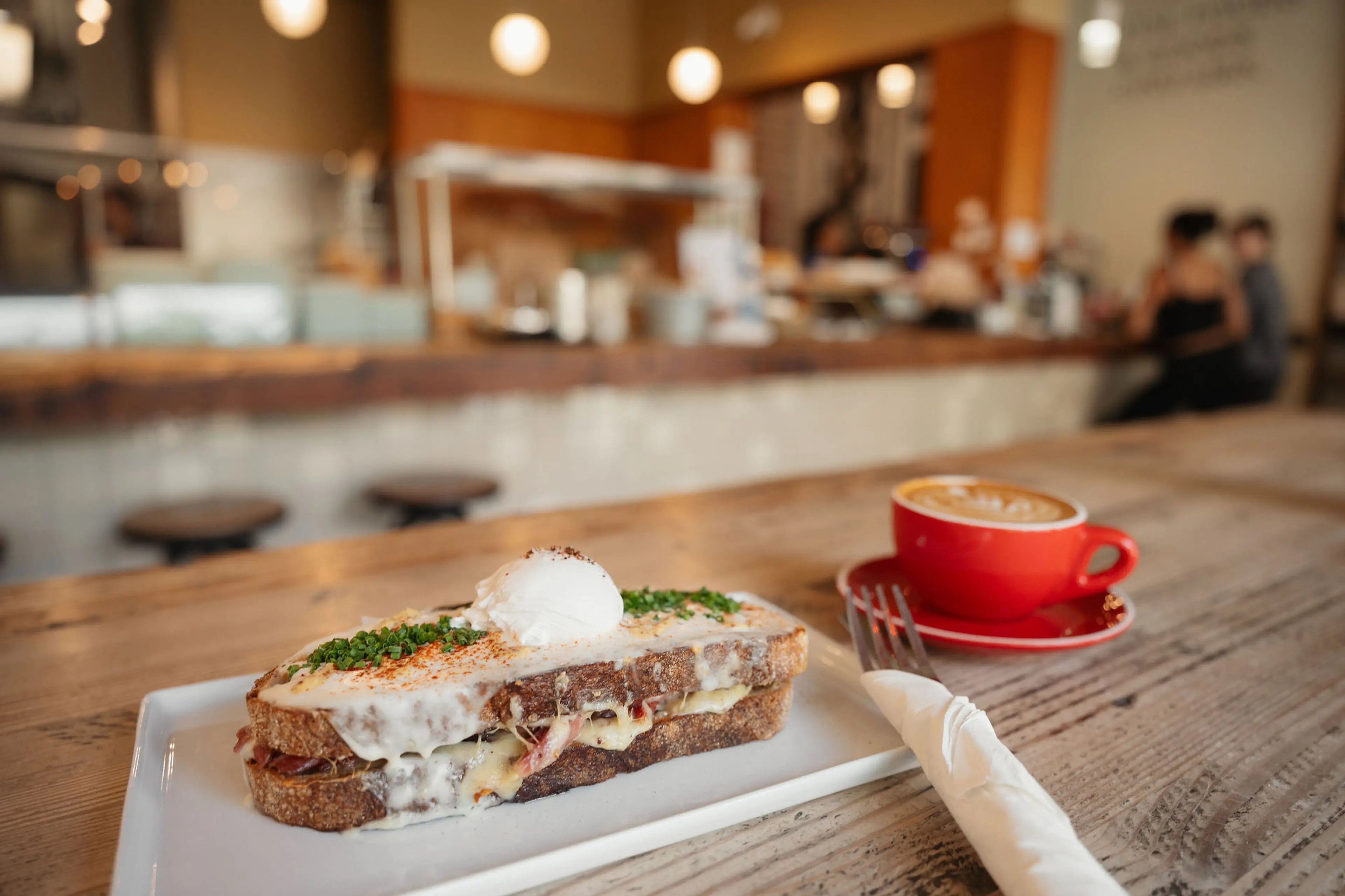 Toasted sandwich with melted cheese and herbs topped with a poached egg on a white plate, next to a red cup of coffee on a wooden table in a cafe.