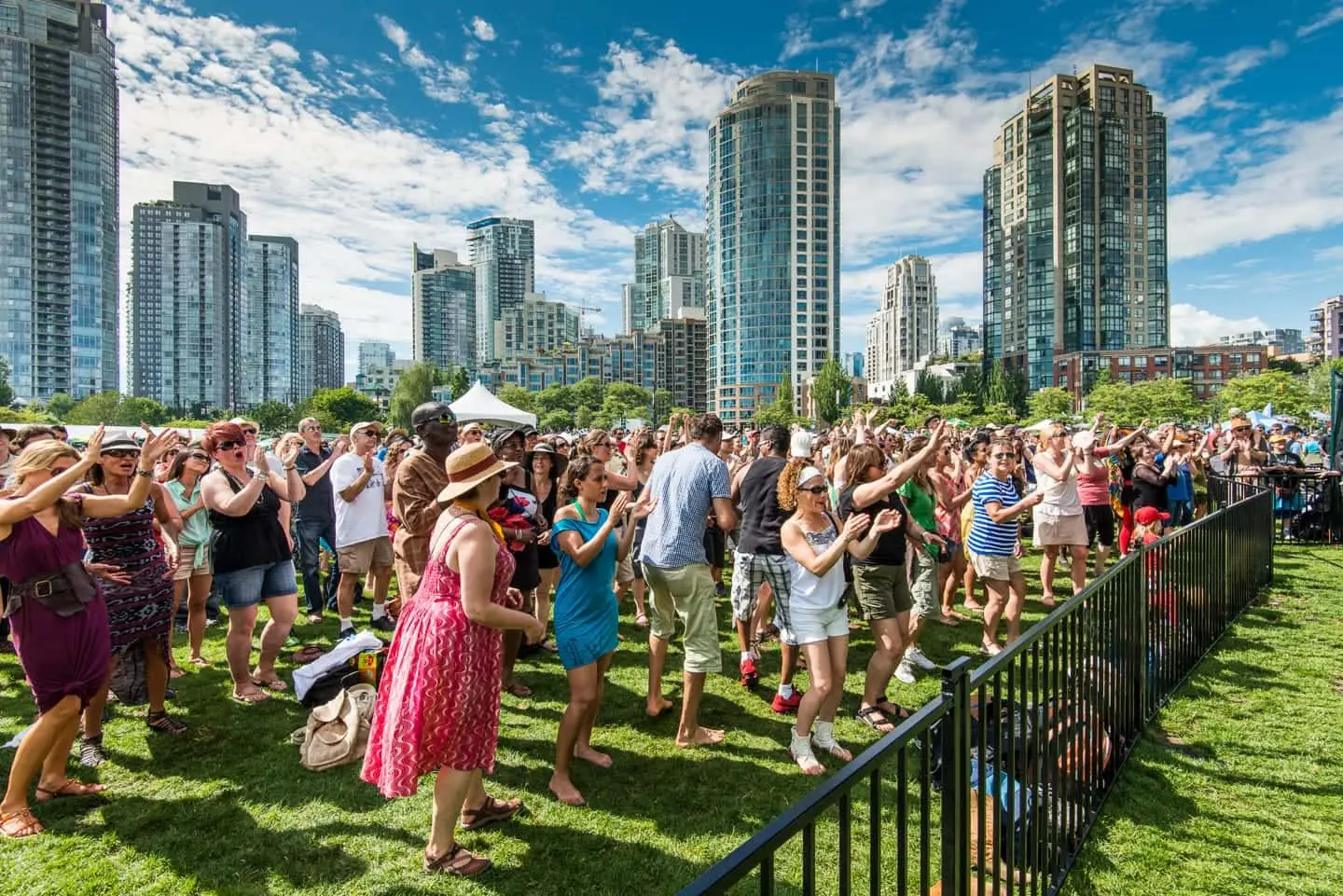 a large crowd of people enjoying a sunny day on a grassy field, with modern high-rise buildings in the background.
