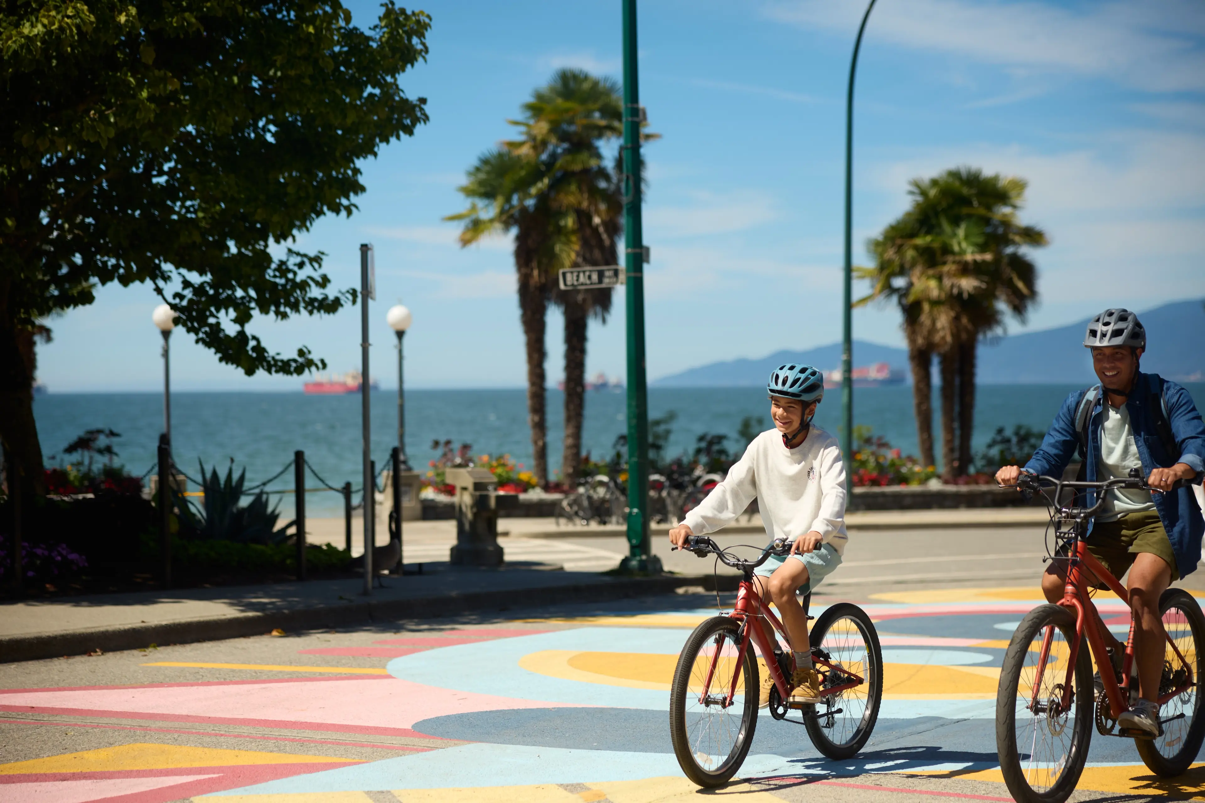 Two people wearing helmets riding bicycles on a colorful street near the beach with palm trees and ocean in the background.
