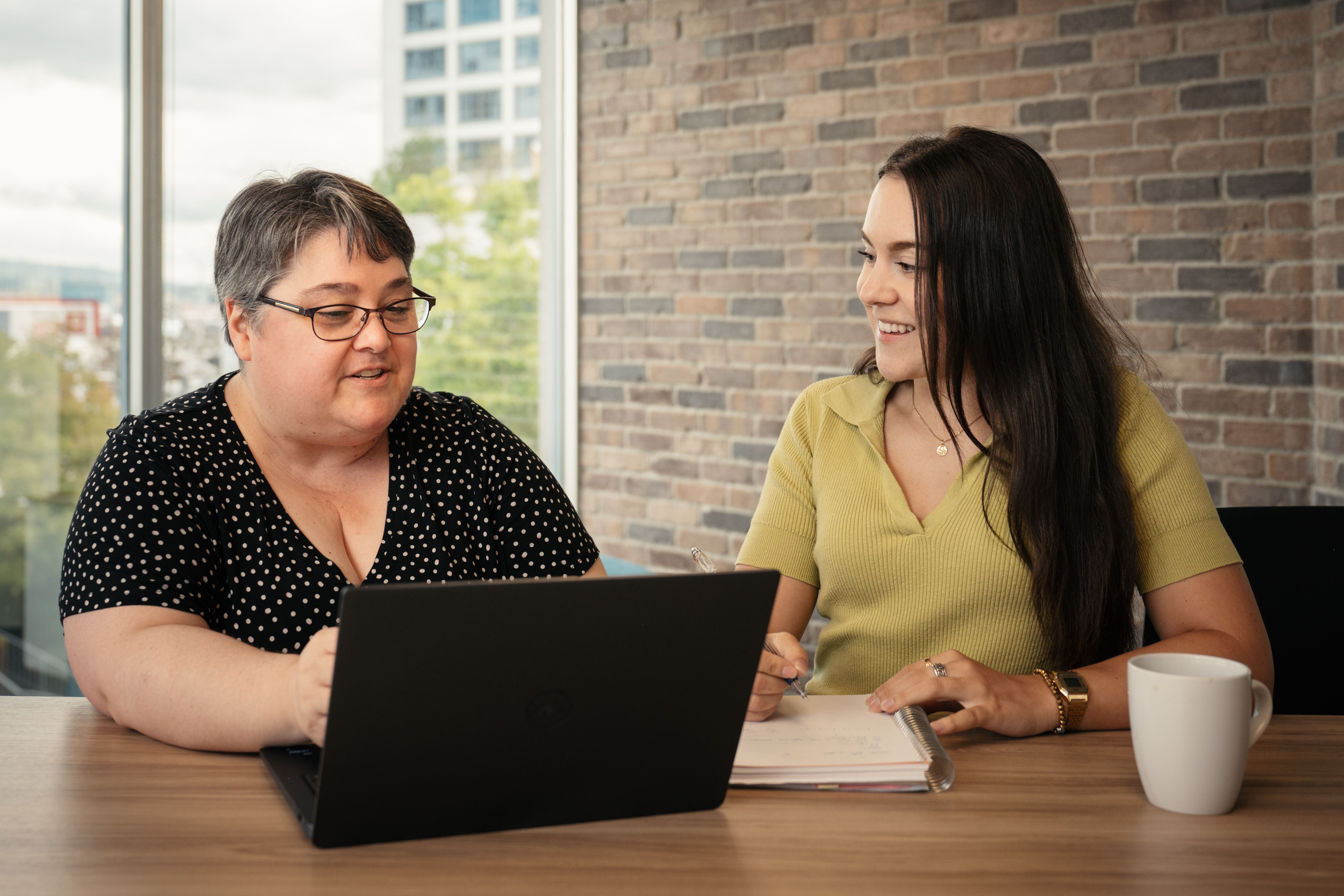 Two women sitting at a table with a laptop and notebook, discussing work in an office with a brick wall background.