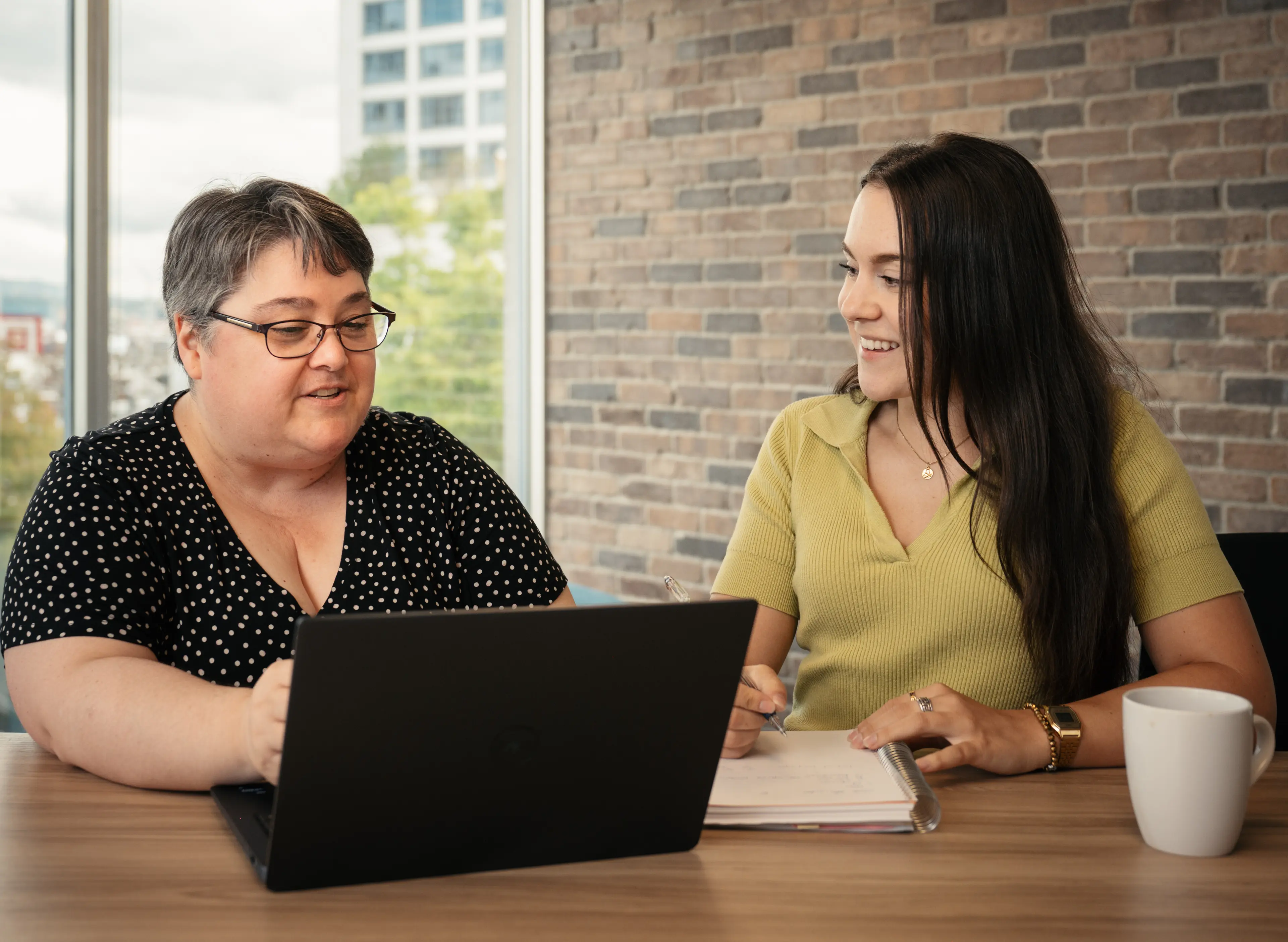 Two women sitting at a table with a laptop and notebook, discussing work in an office with a brick wall background.