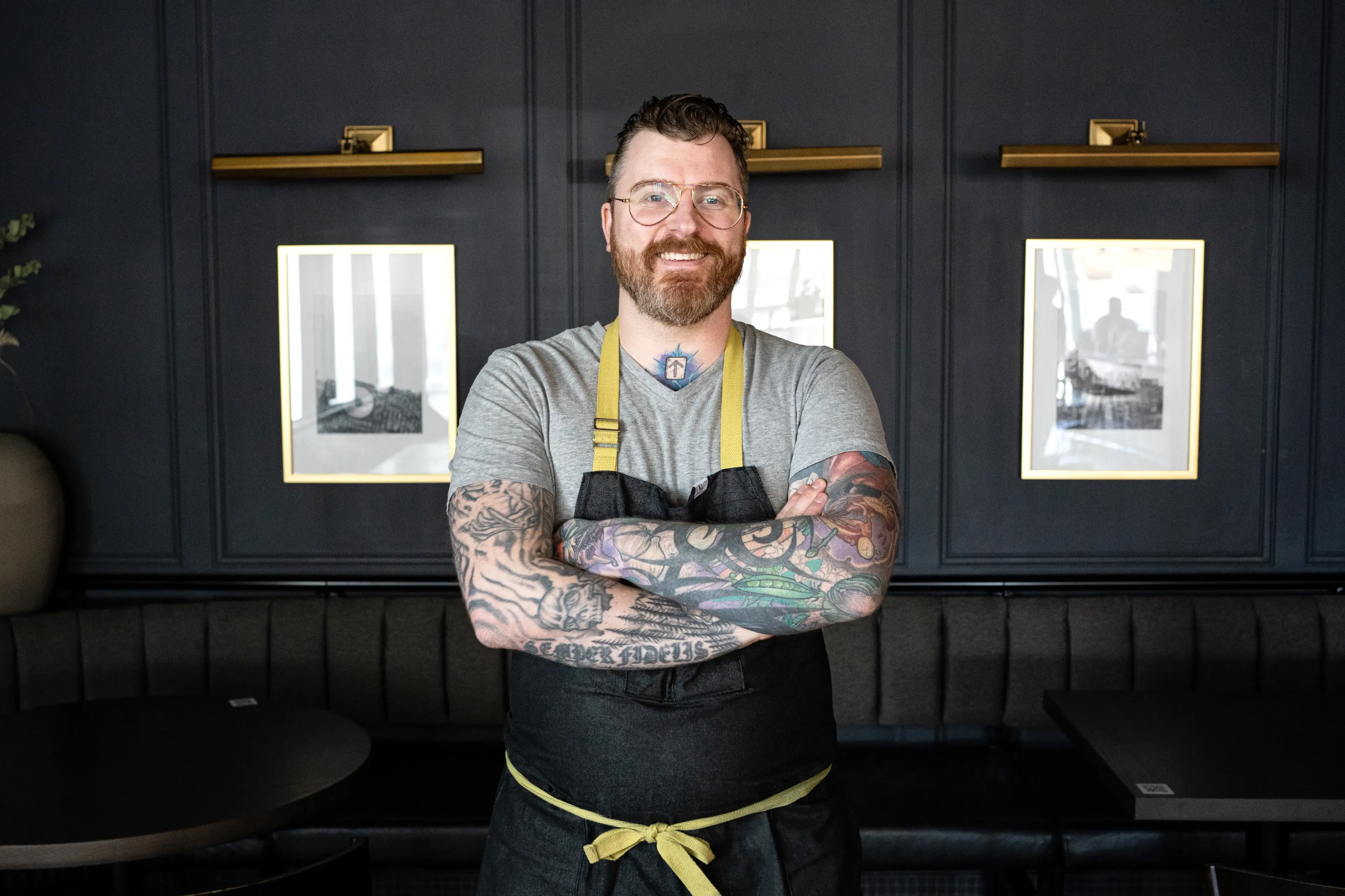 Person with glasses and tattoos wearing a gray shirt and black apron standing with arms crossed in a dark room with framed pictures on the wall.