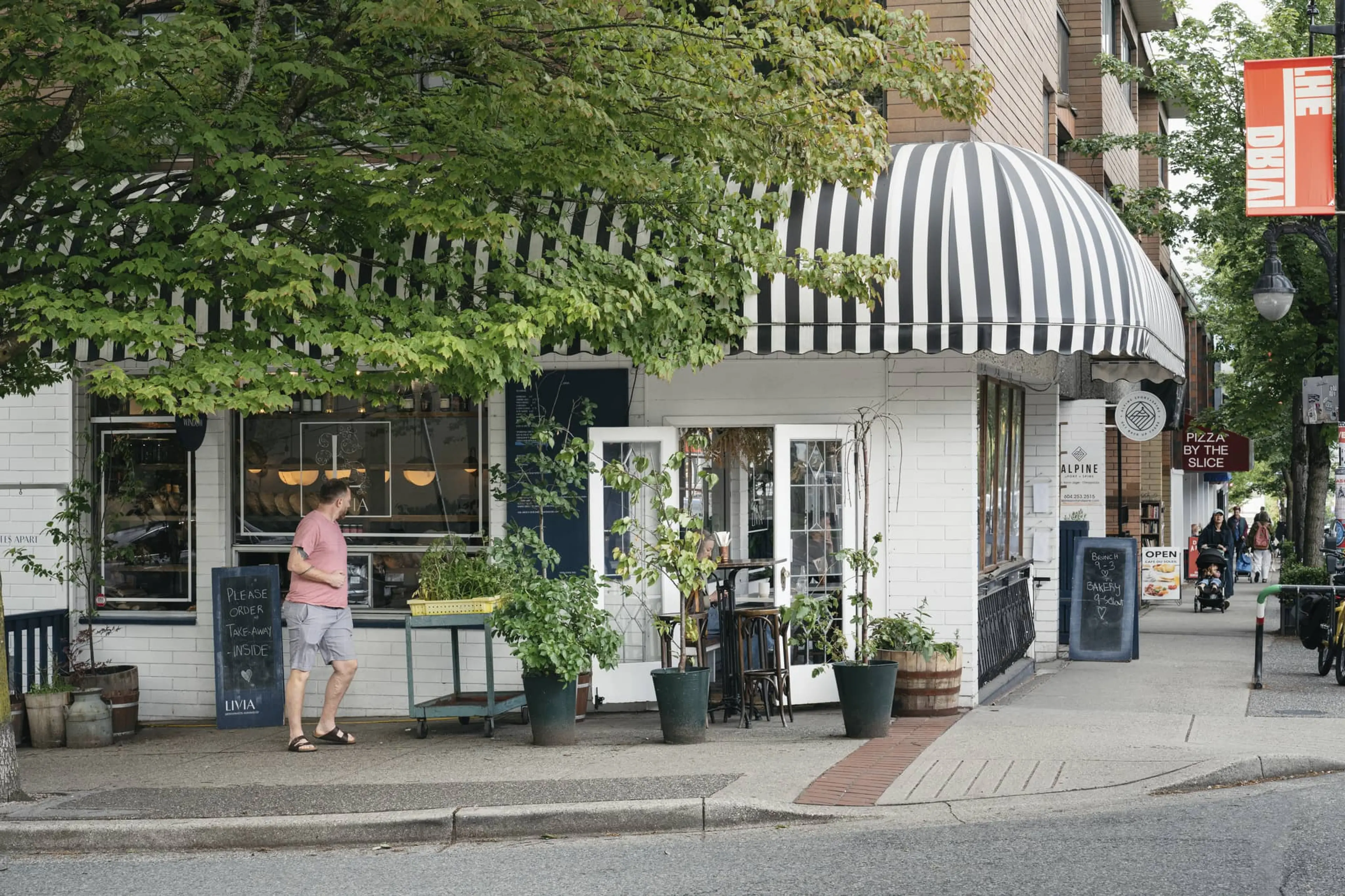 Man in pink shirt and shorts walking past a cafe with black and white striped awning and potted plants outside.
