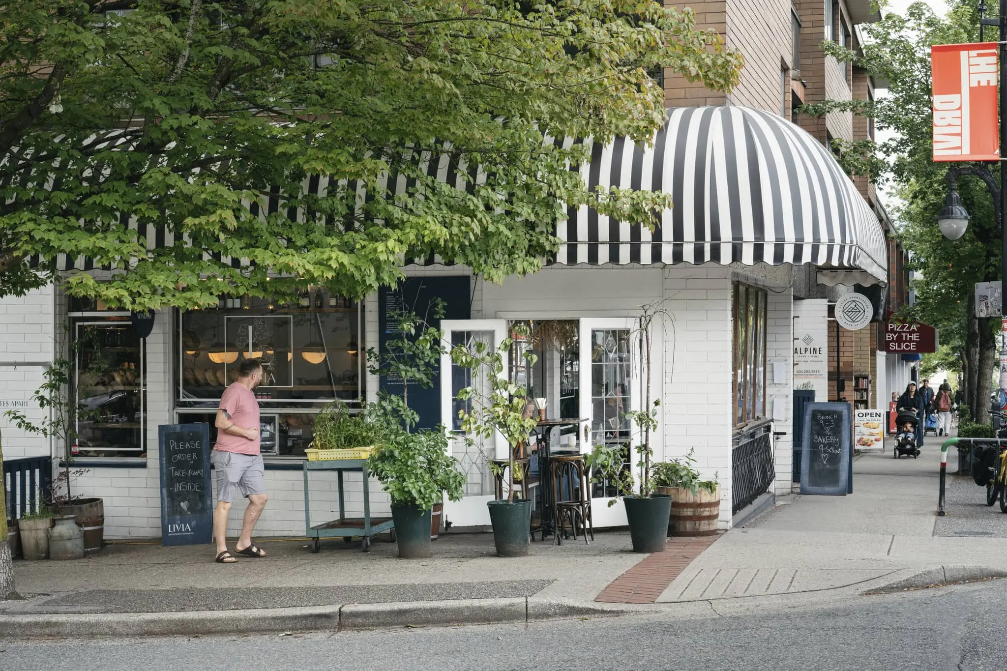 Man in pink shirt and shorts walking past a cafe with black and white striped awning and potted plants outside.