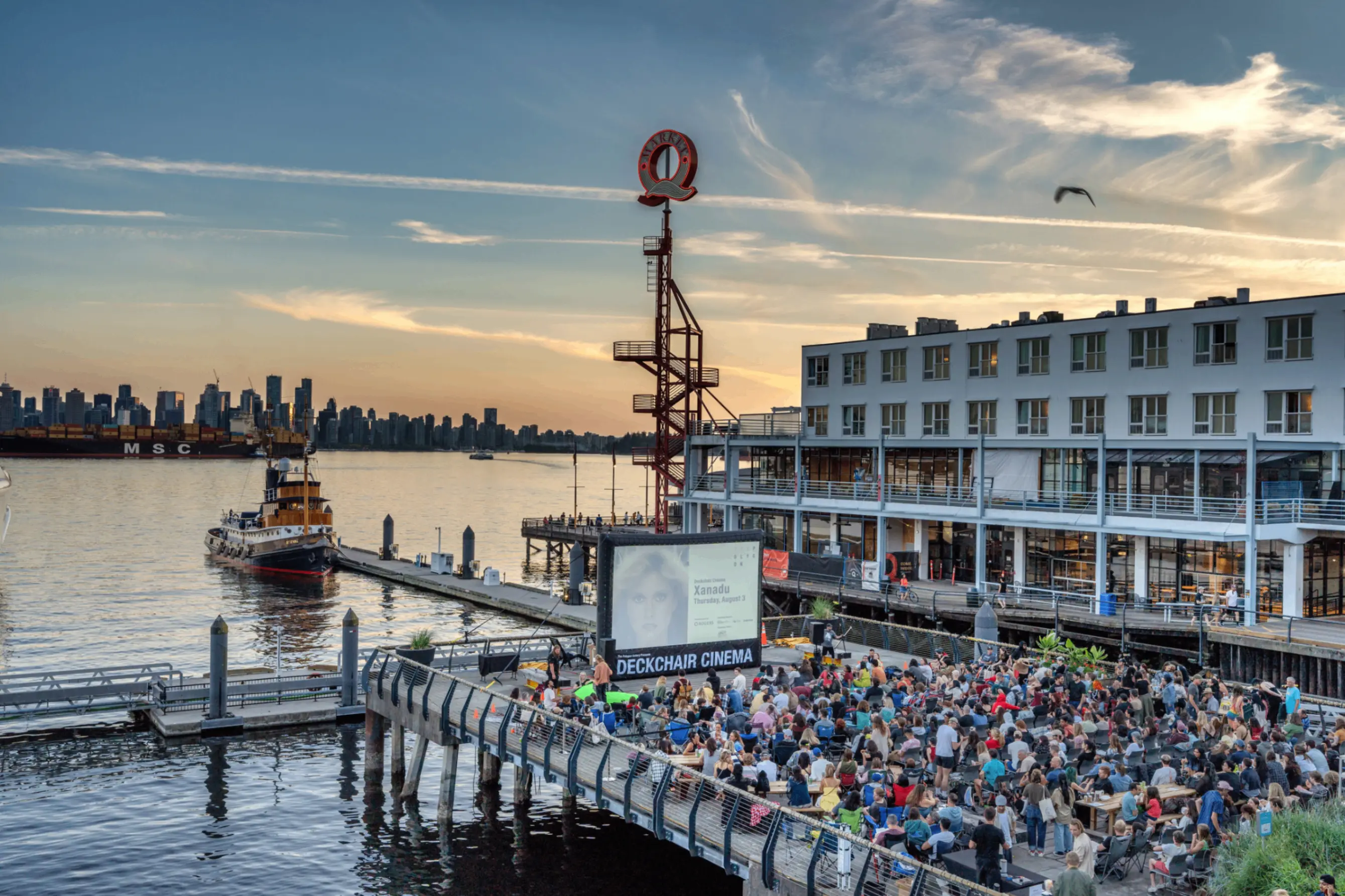 Crowd gathered at outdoor Deckchair Cinema on a pier by waterfront with city skyline and boat in background at sunset.
