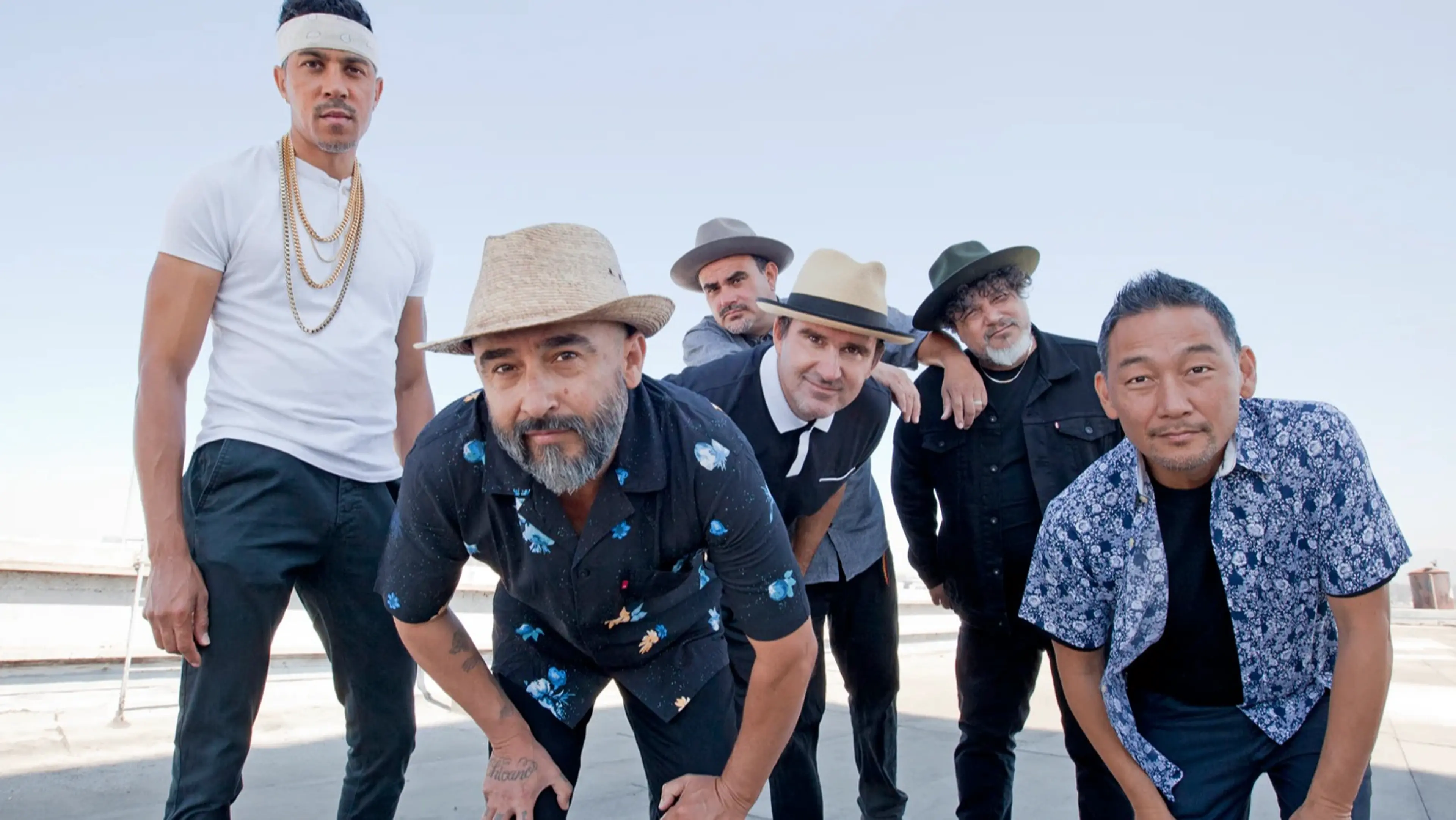 Six people posing outdoors against a clear sky, wearing hats and casual clothing, some leaning forward.