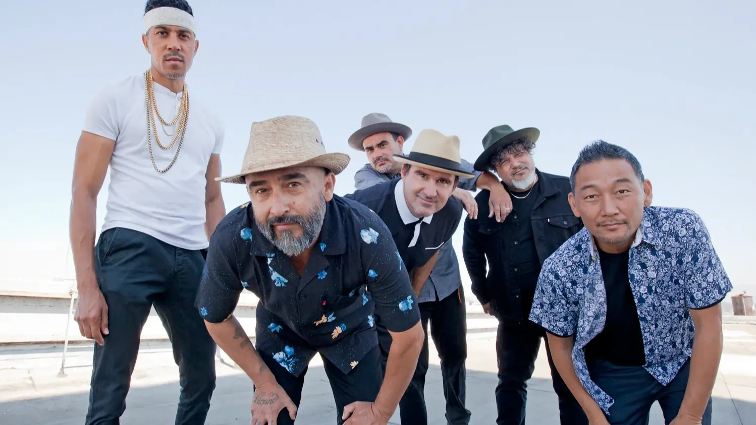 Six people posing outdoors against a clear sky, wearing hats and casual clothing, some leaning forward.