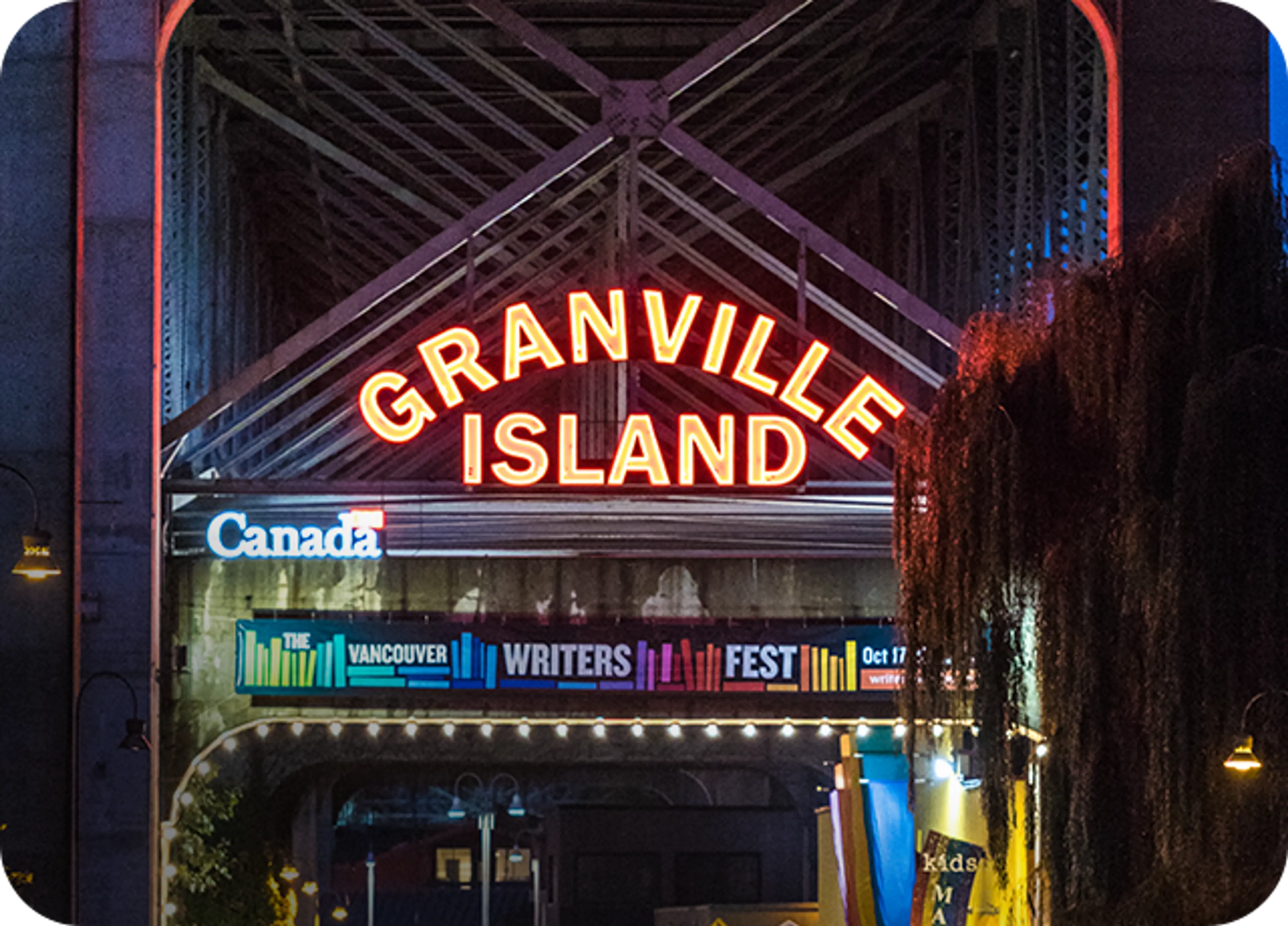Neon sign reading Granville Island above a banner for the Vancouver Writers Fest under a bridge at night.