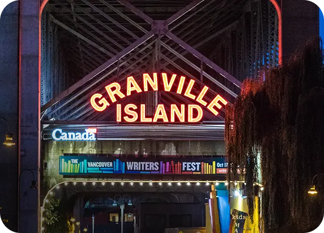 Neon sign reading Granville Island above a banner for the Vancouver Writers Fest under a bridge at night.