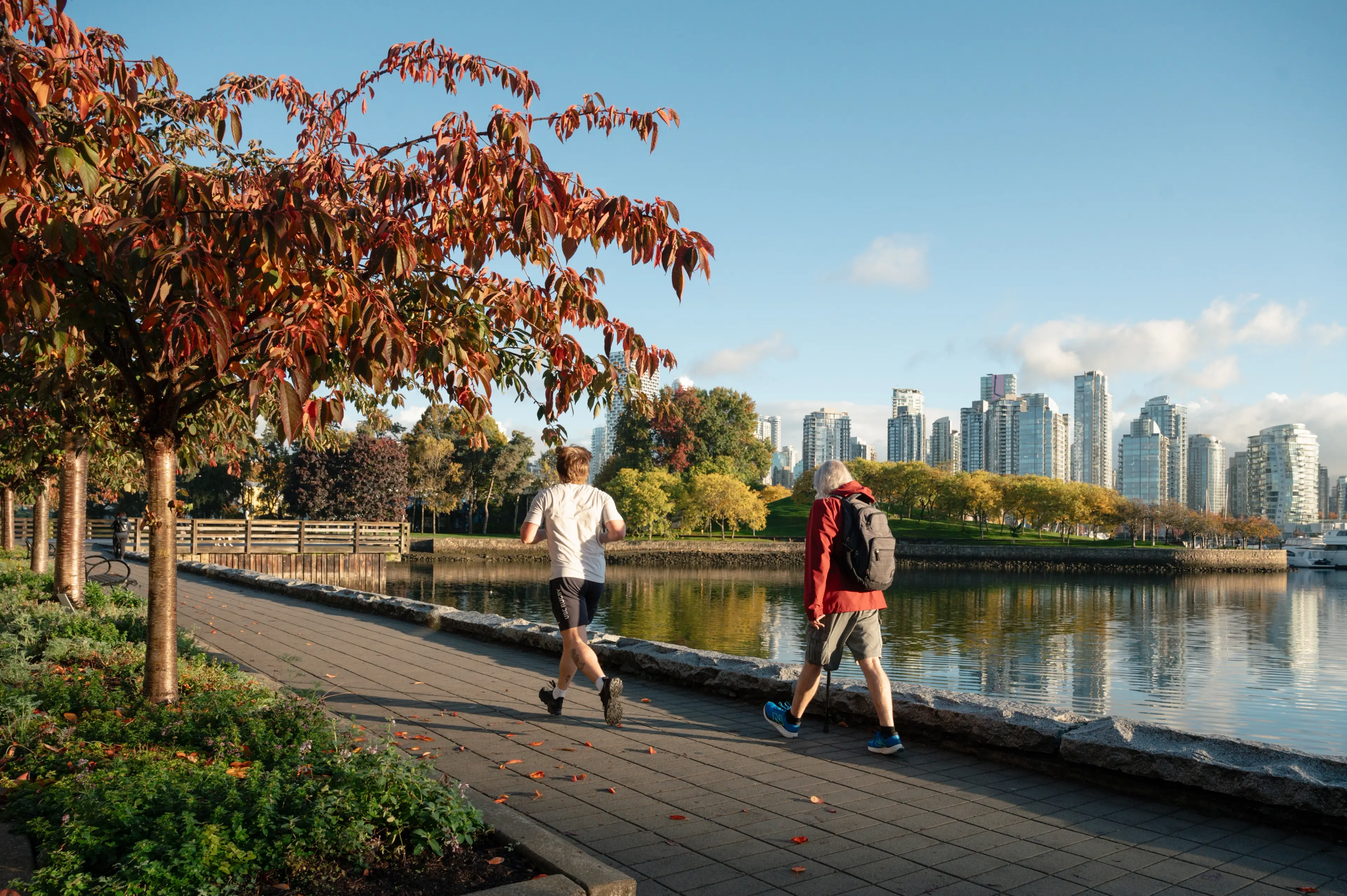 Two people walking and jogging on a paved path beside a body of water with city buildings and autumn trees in the background.