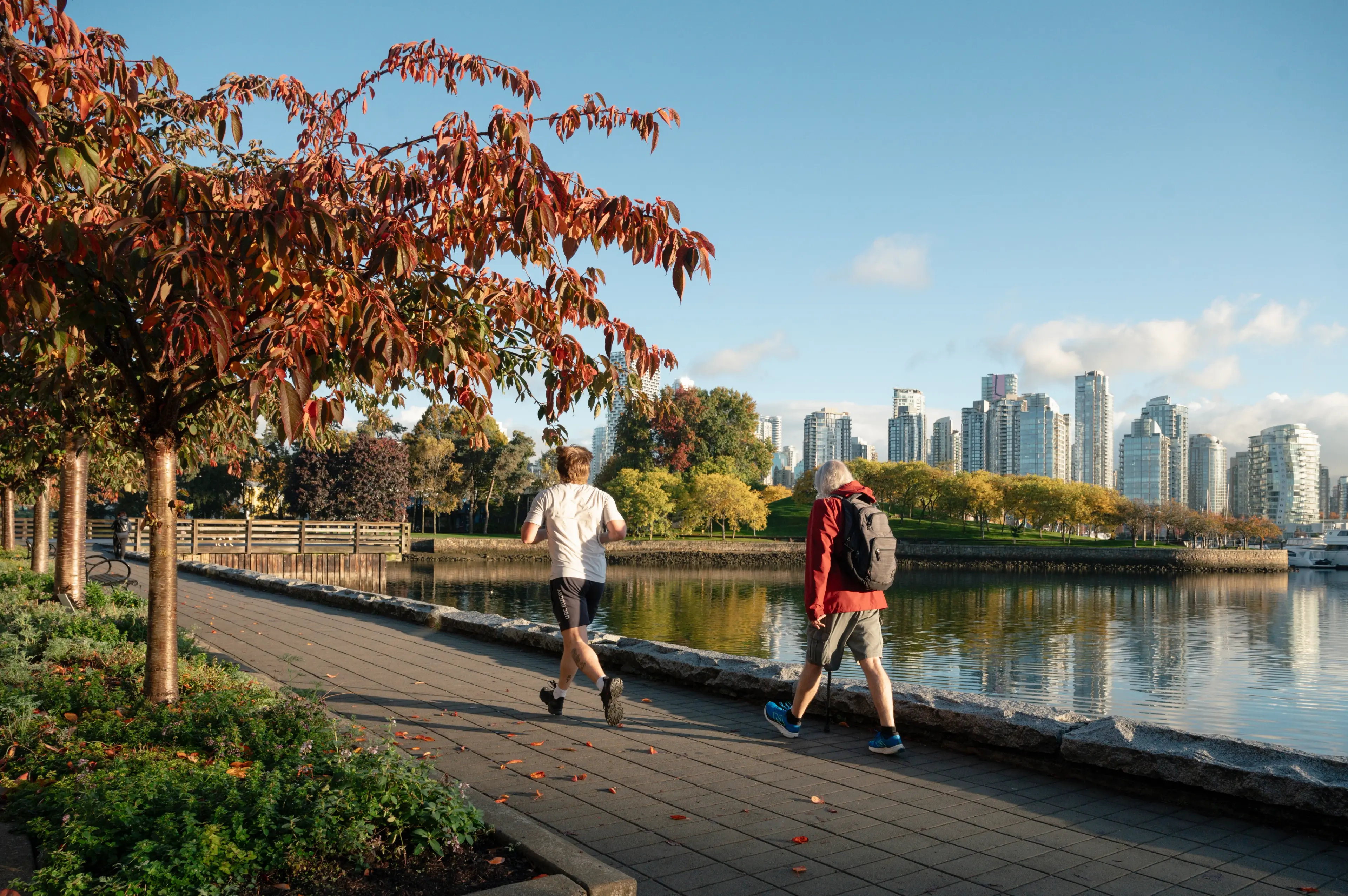 Two people walking and jogging on a paved path beside a body of water with city buildings and autumn trees in the background.
