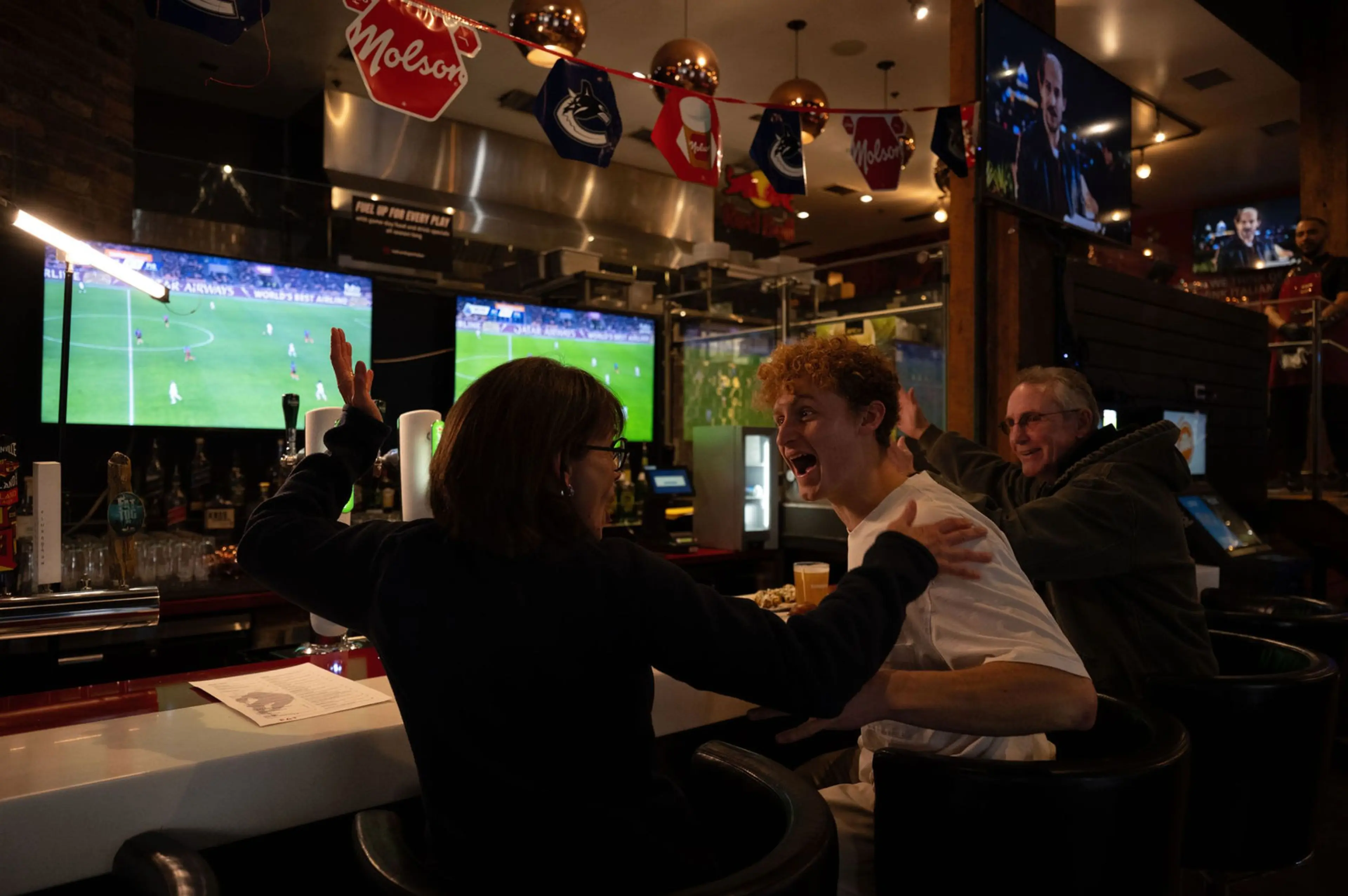 Three people at a bar watching a soccer game on TV, two are excitedly reacting with raised arms and smiles.