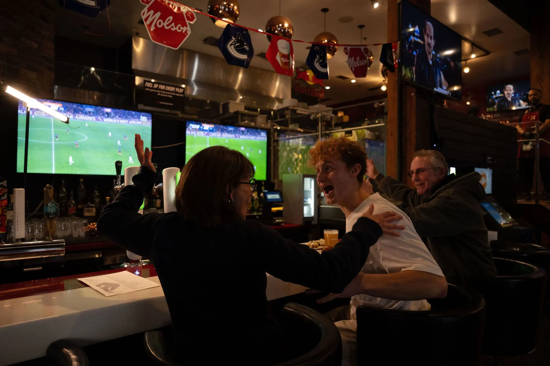 Three people at a bar watching a soccer game on TV, two are excitedly reacting with raised arms and smiles.