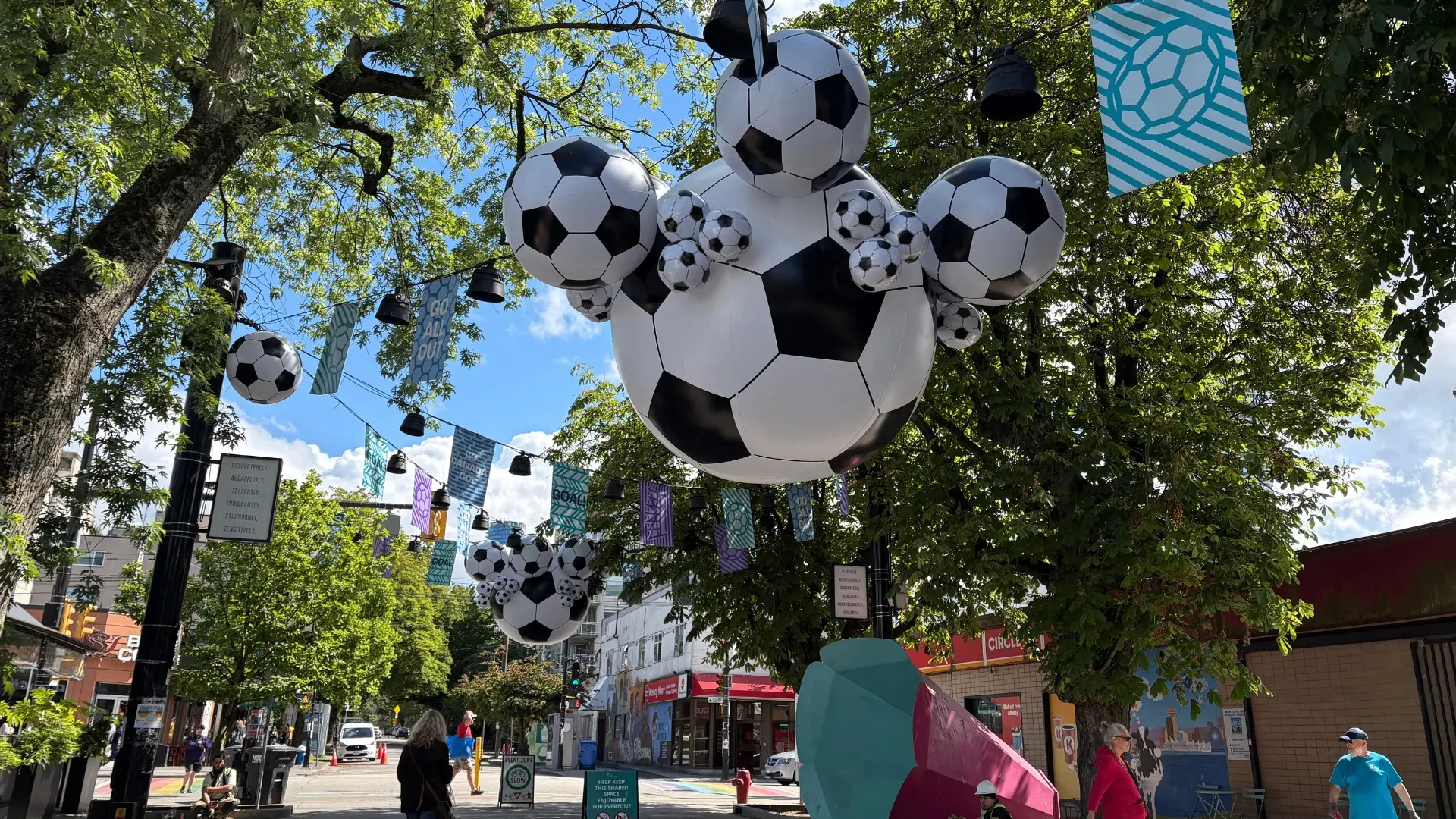 Large and small soccer ball decorations hanging from trees and streetlights on a sunny urban street.