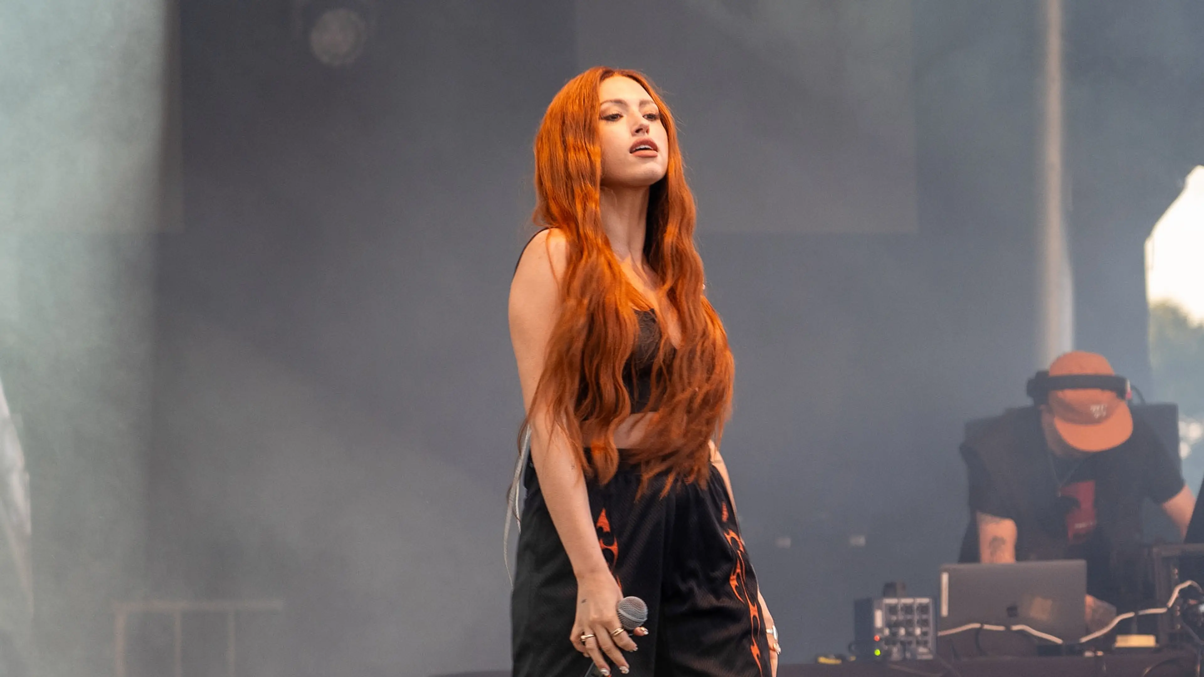 Person with long red hair holding a microphone on stage with a DJ in the background.