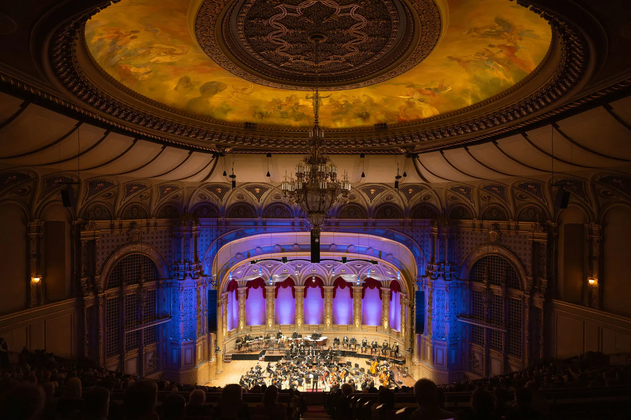 Orchestra performing on stage in a grand concert hall with ornate ceiling and chandelier, audience seated in foreground.