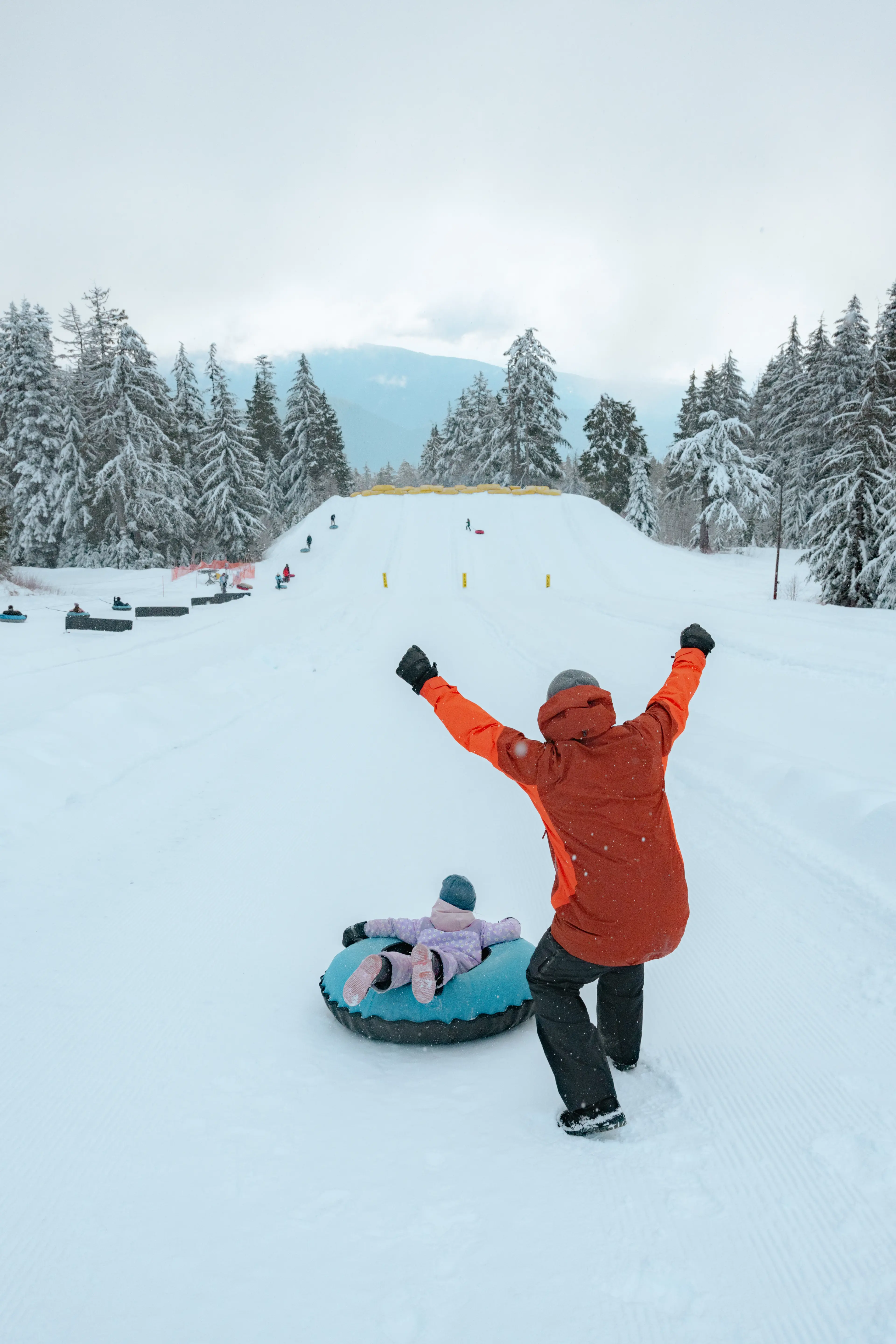 Person in orange jacket cheering next to child lying on blue snow tube on snowy hill with snow-covered trees.