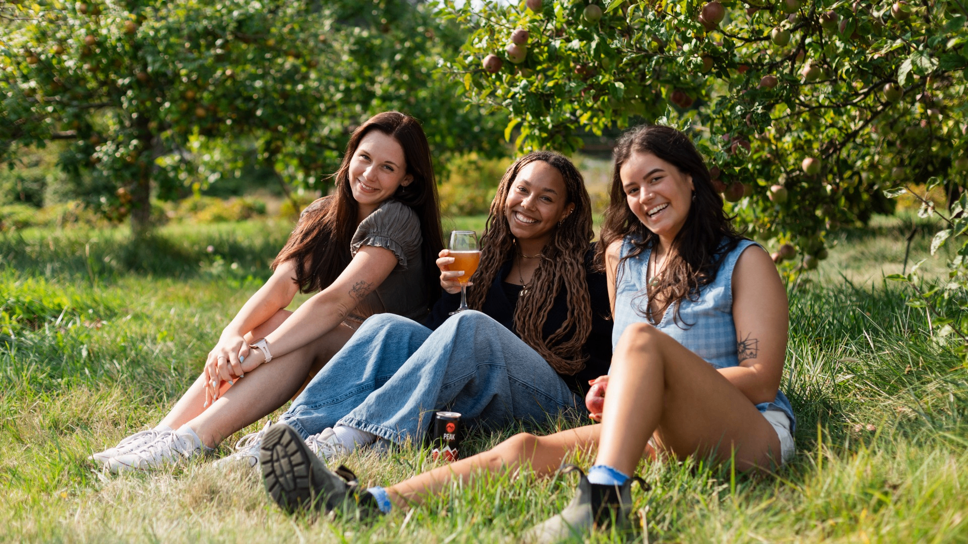Three people sitting on grass under apple trees, smiling, one holding a drink and another holding an apple.