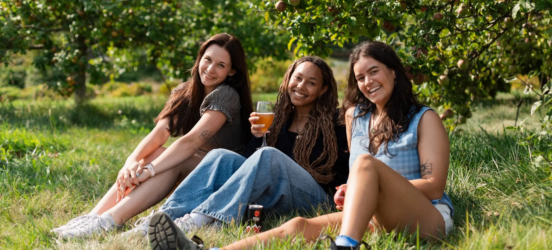 Three people sitting on grass under apple trees, smiling, one holding a drink and another holding an apple.