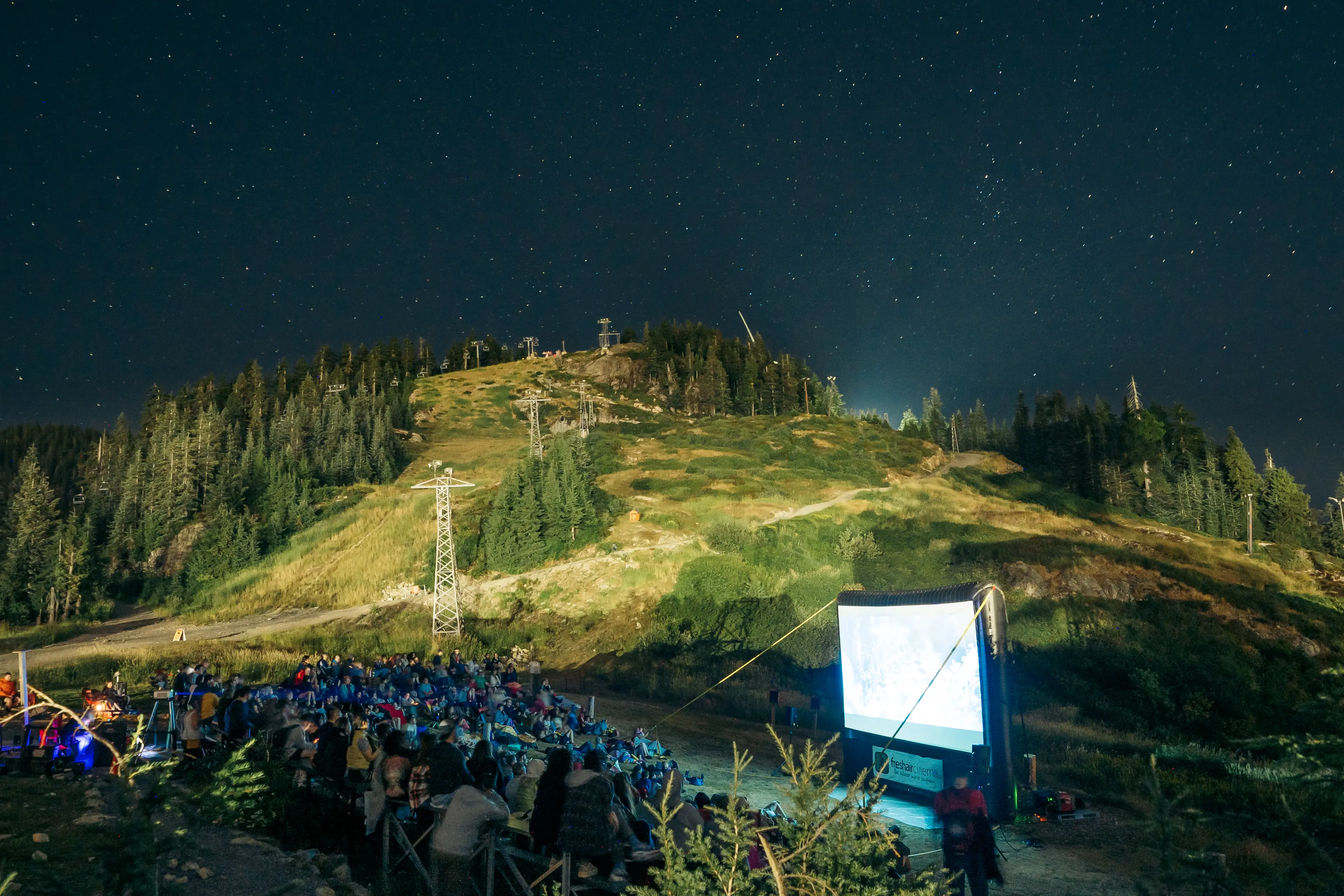 Outdoor movie screening at night with a crowd seated facing a large screen in a mountainous area under a starry sky.