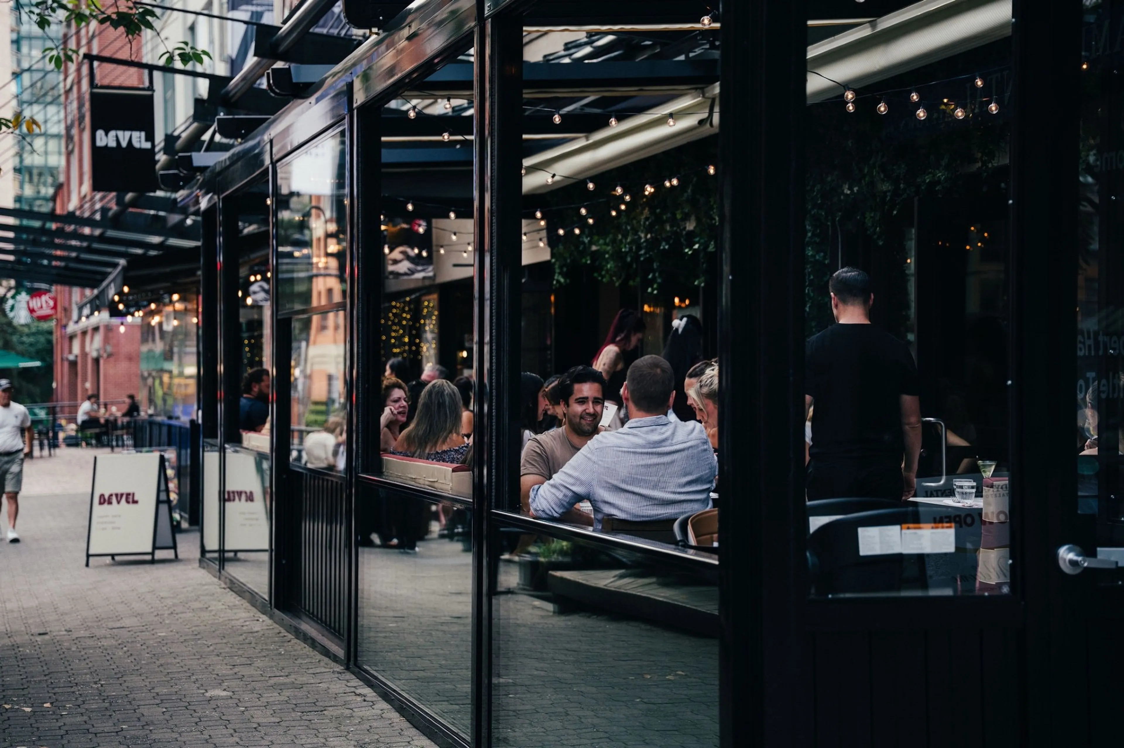 People sitting and talking inside a glass-walled restaurant with string lights, and a person walking on the sidewalk outside.