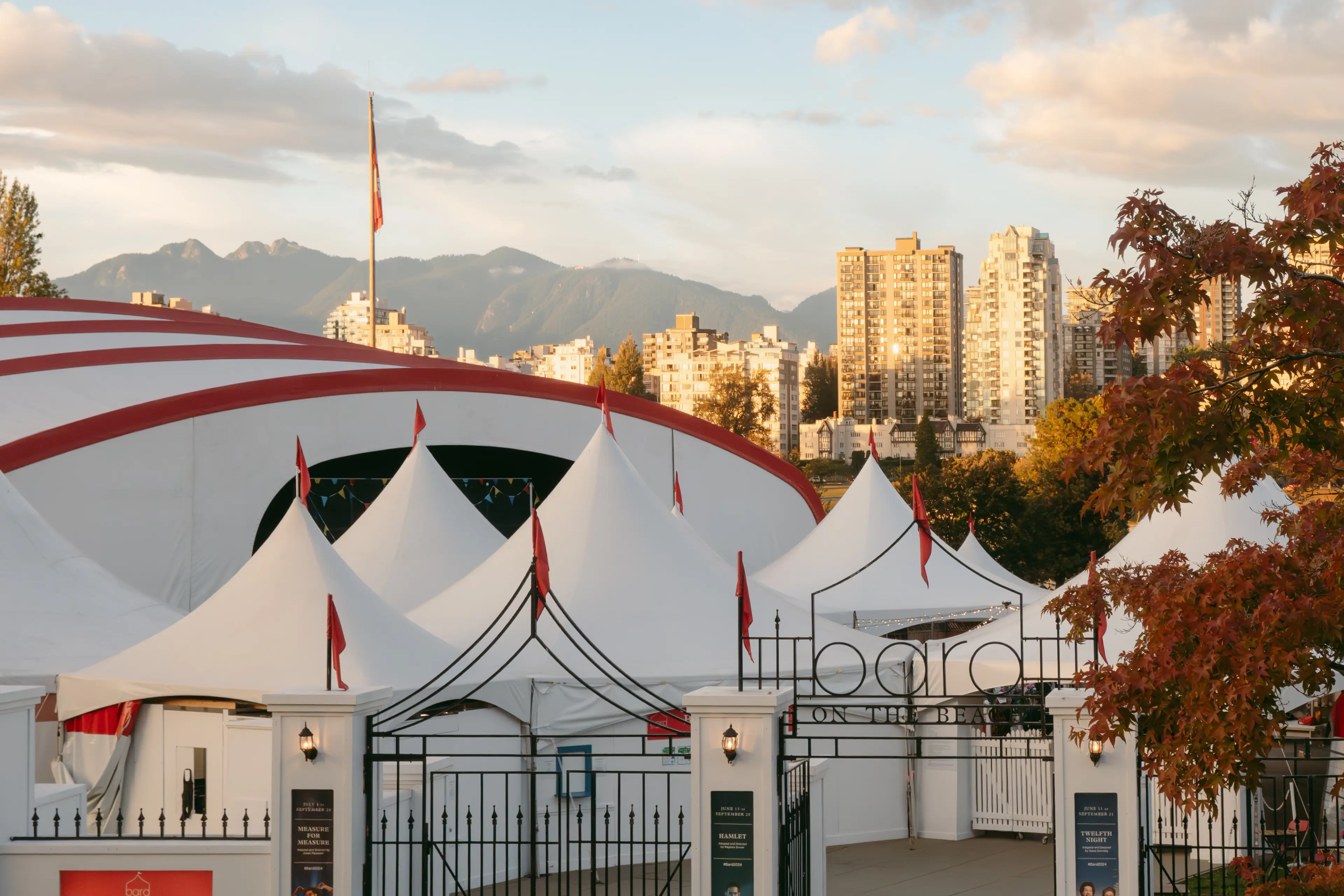 White tents with red flags and a large white and red circus tent in front of city buildings and mountains under a cloudy sky.