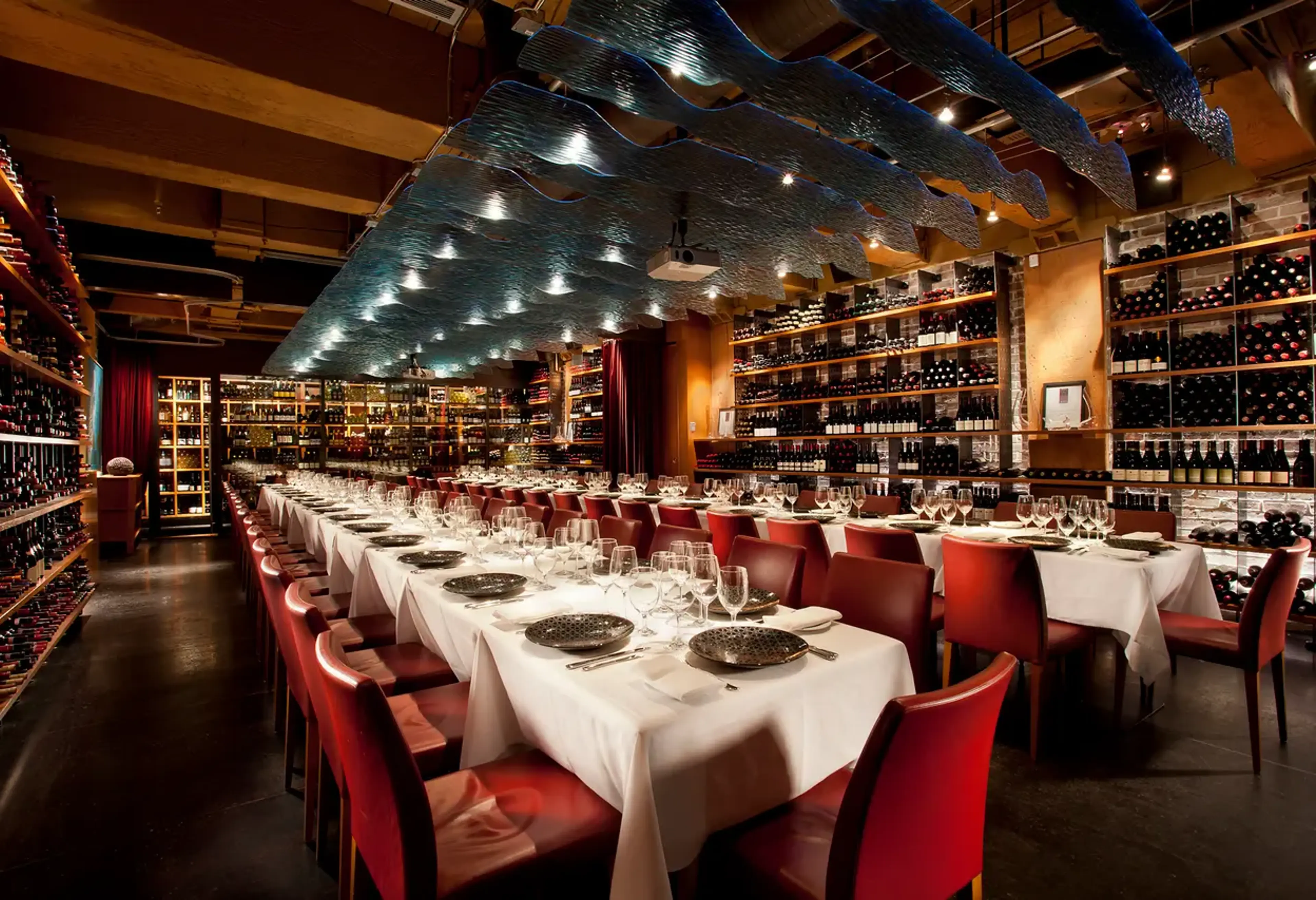 Long dining tables with white tablecloths and red chairs set with plates and glasses in a wine cellar with shelves of wine bottles.